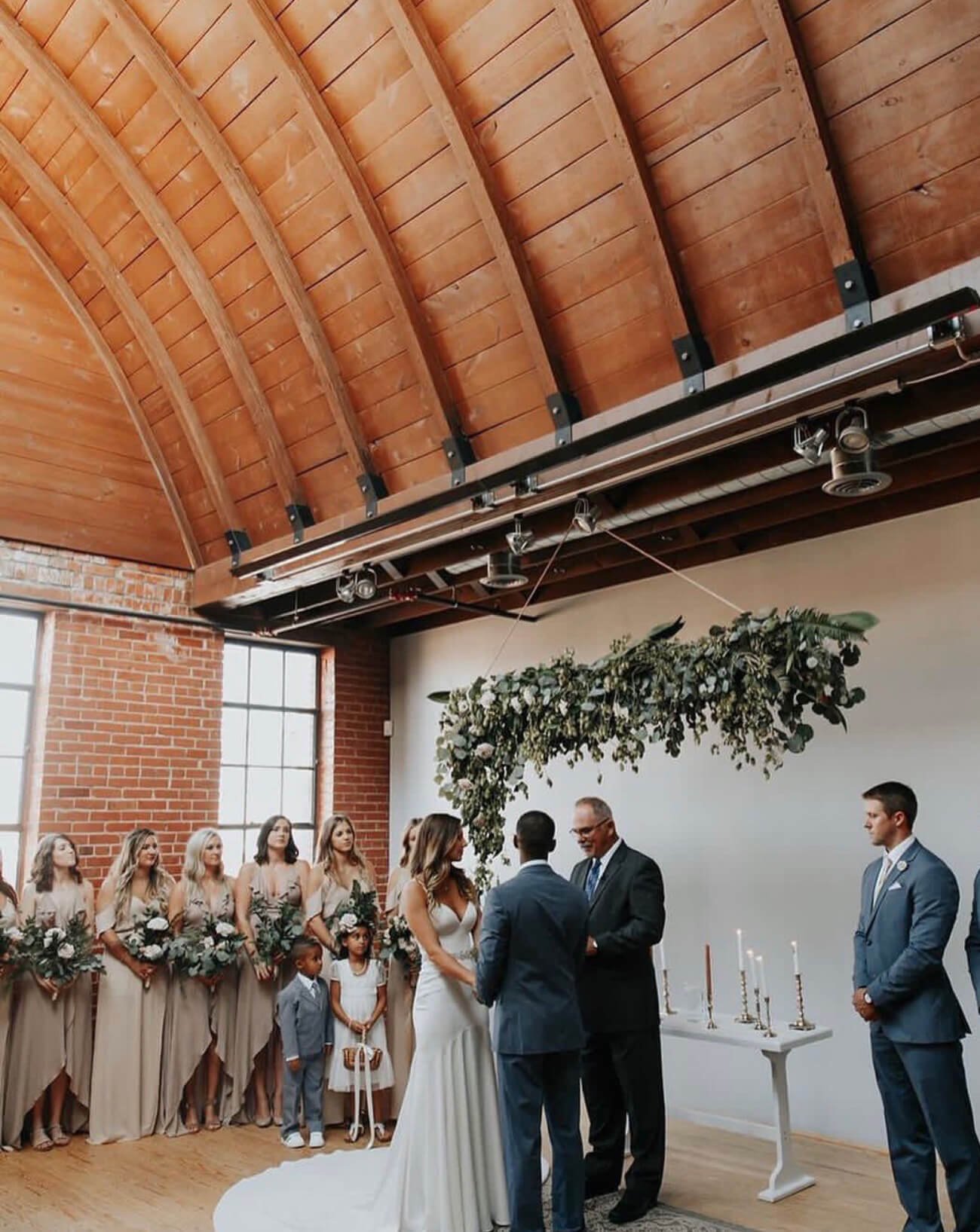 Wedding ceremony at Holloway House beneath a vaulted wood ceiling with exposed brick and greenery installation