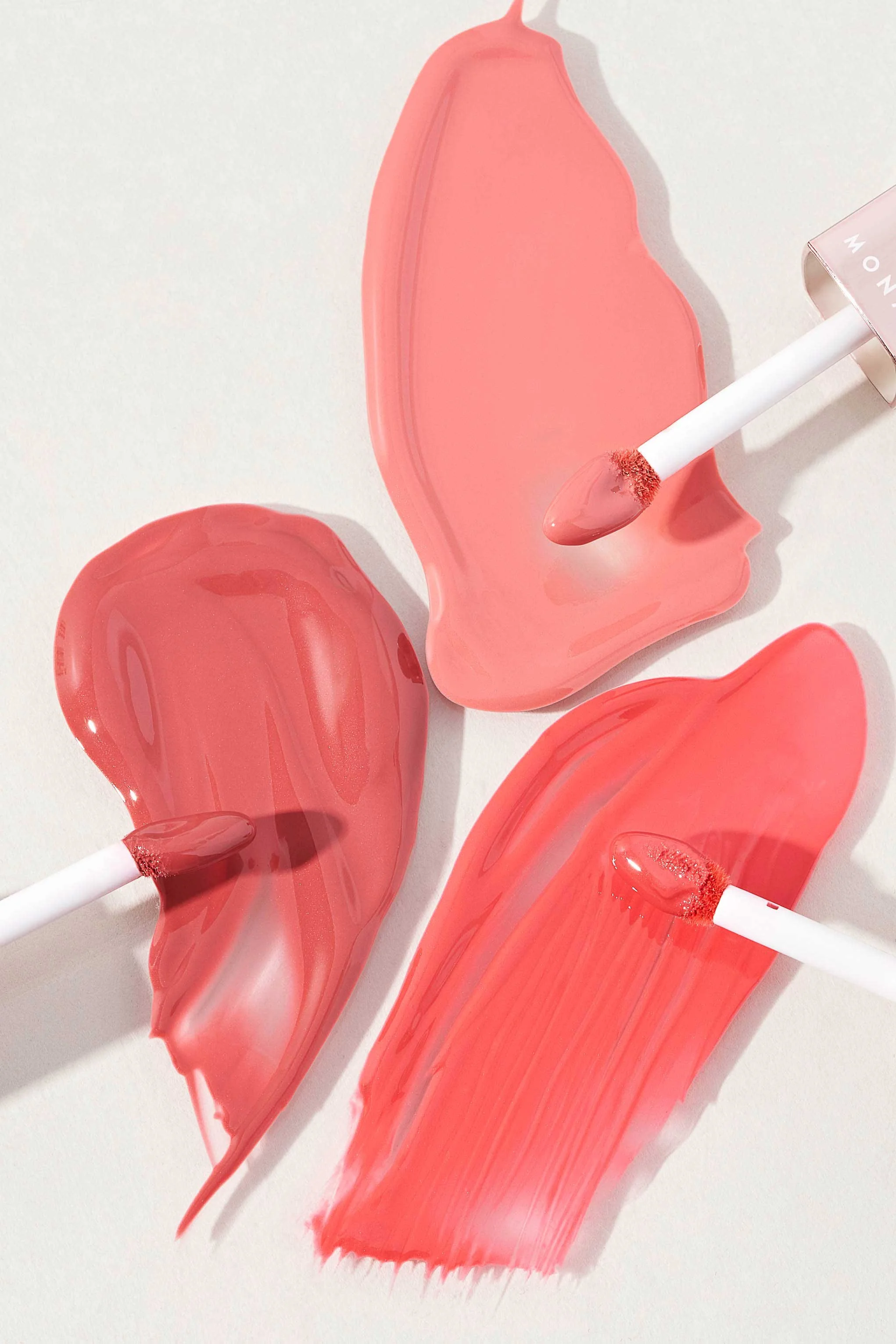 Three smears of pink lipstick with white applicator sticks on white background.