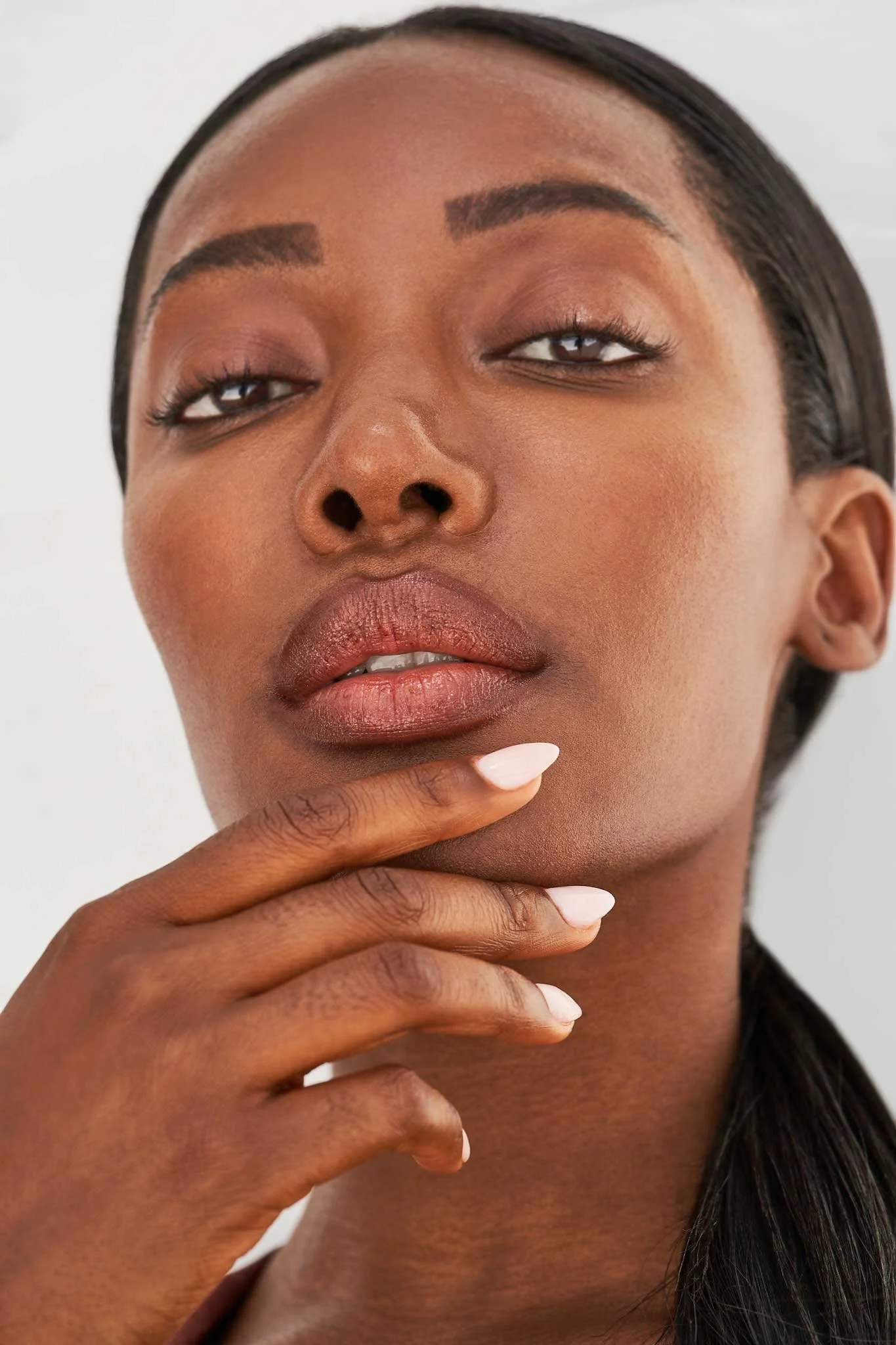 Close-up of a woman’s face with natural makeup, touching her chin with her fingers, dark hair, and a neutral background.