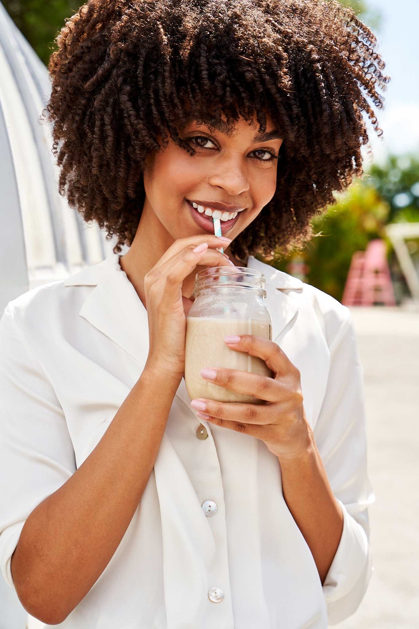 Smiling woman with curly hair drinking a milkshake through a straw outdoors.