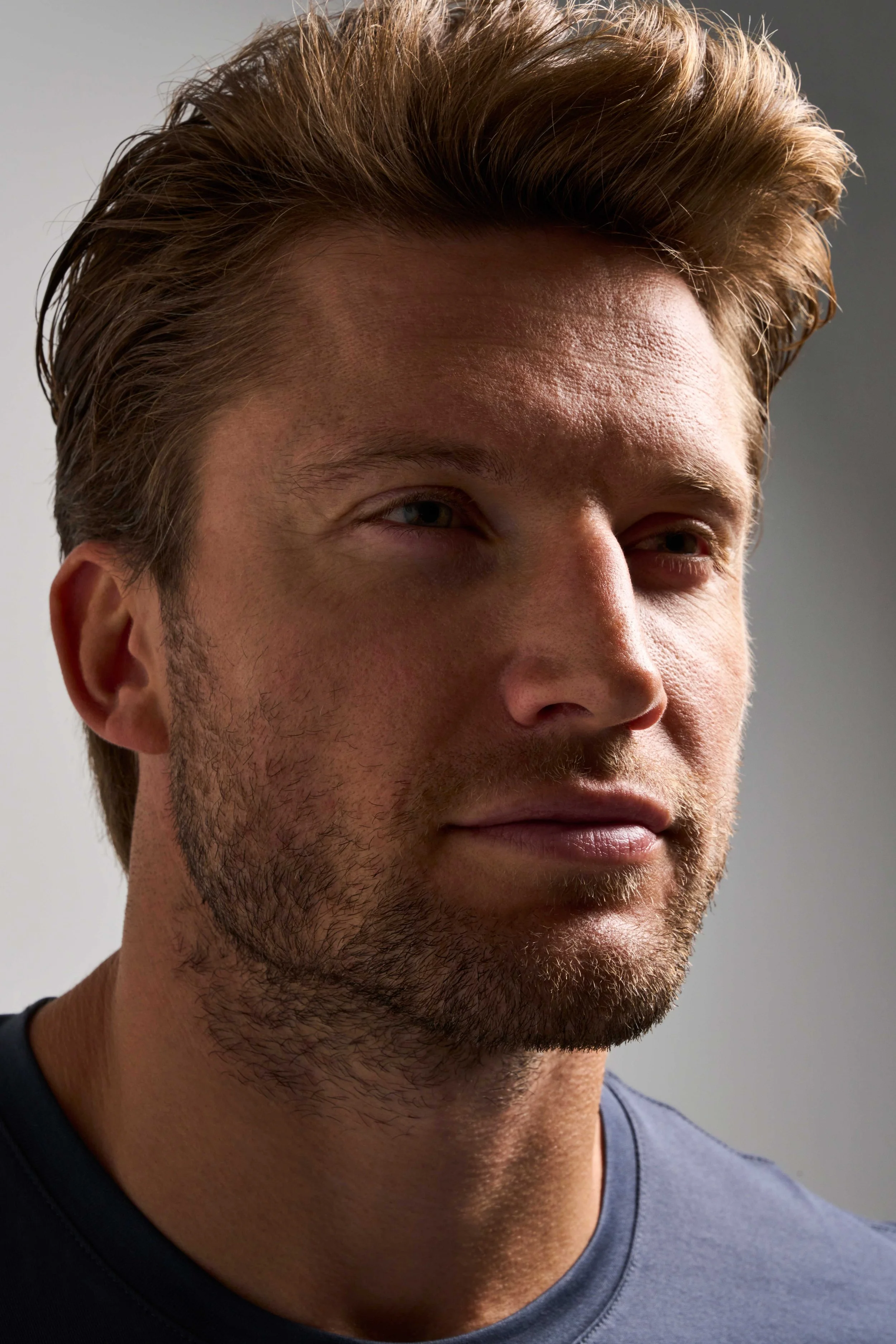 Close-up of a man with brown hair and facial hair, looking slightly to the side against a plain background.