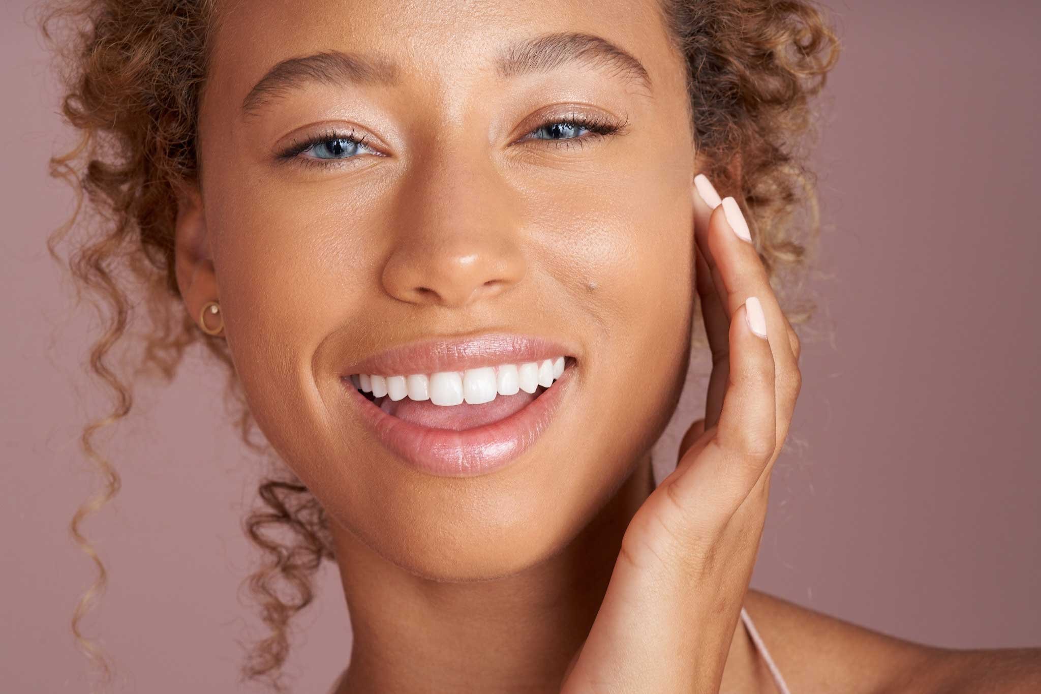 A woman with curly blonde hair smiling and touching her face, with clear skin and bright eyes, against a neutral pink background.