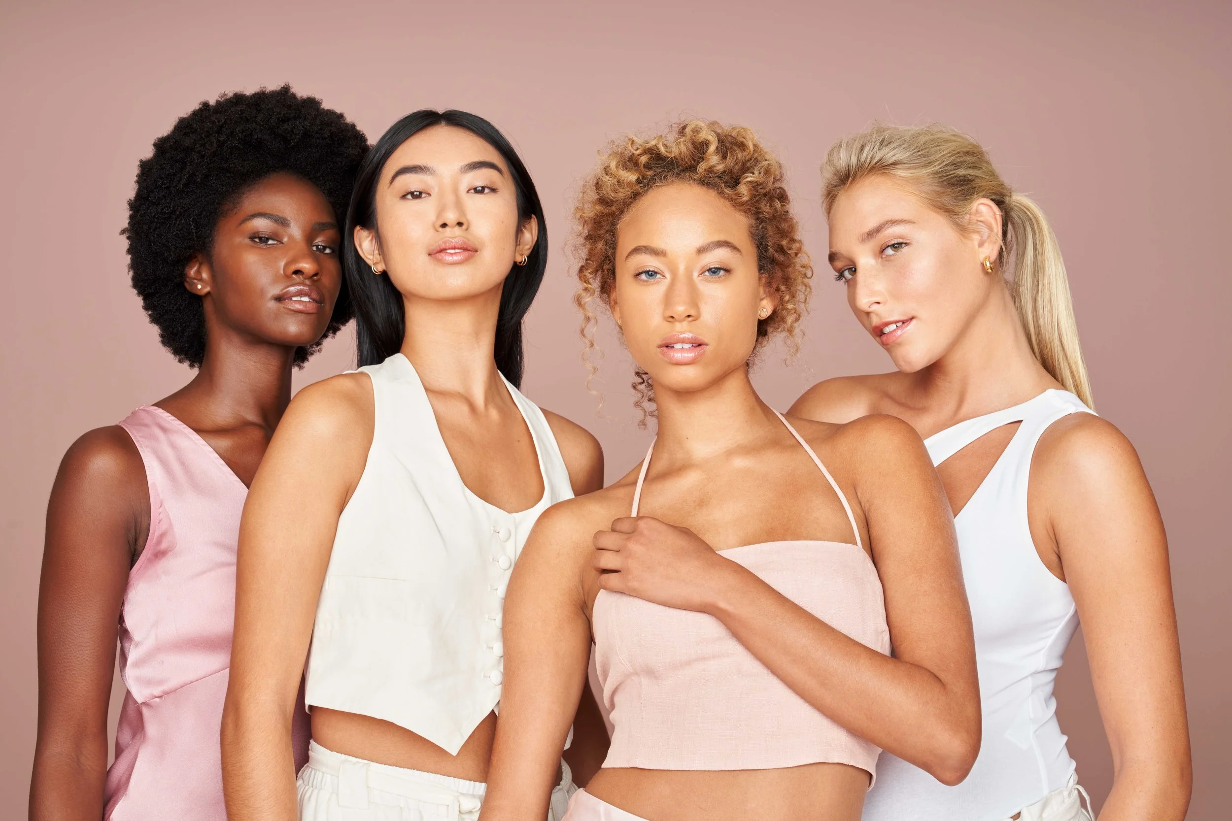 Four women posing together against a plain background, wearing stylish sleeveless tops and looking confidently at the camera.