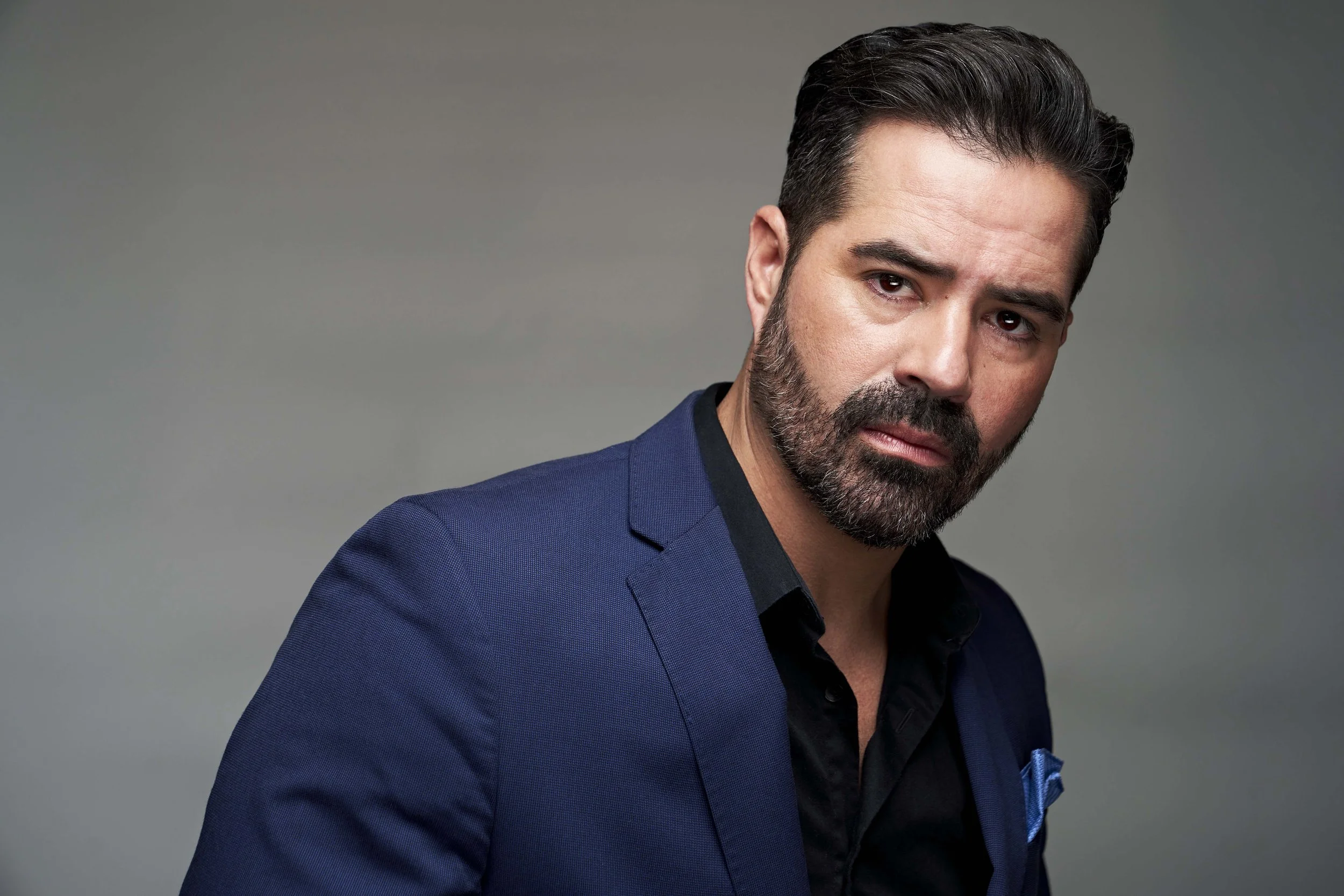 A man with dark hair, a beard, wearing a dark blue suit and black shirt, looking serious against a plain gray background.