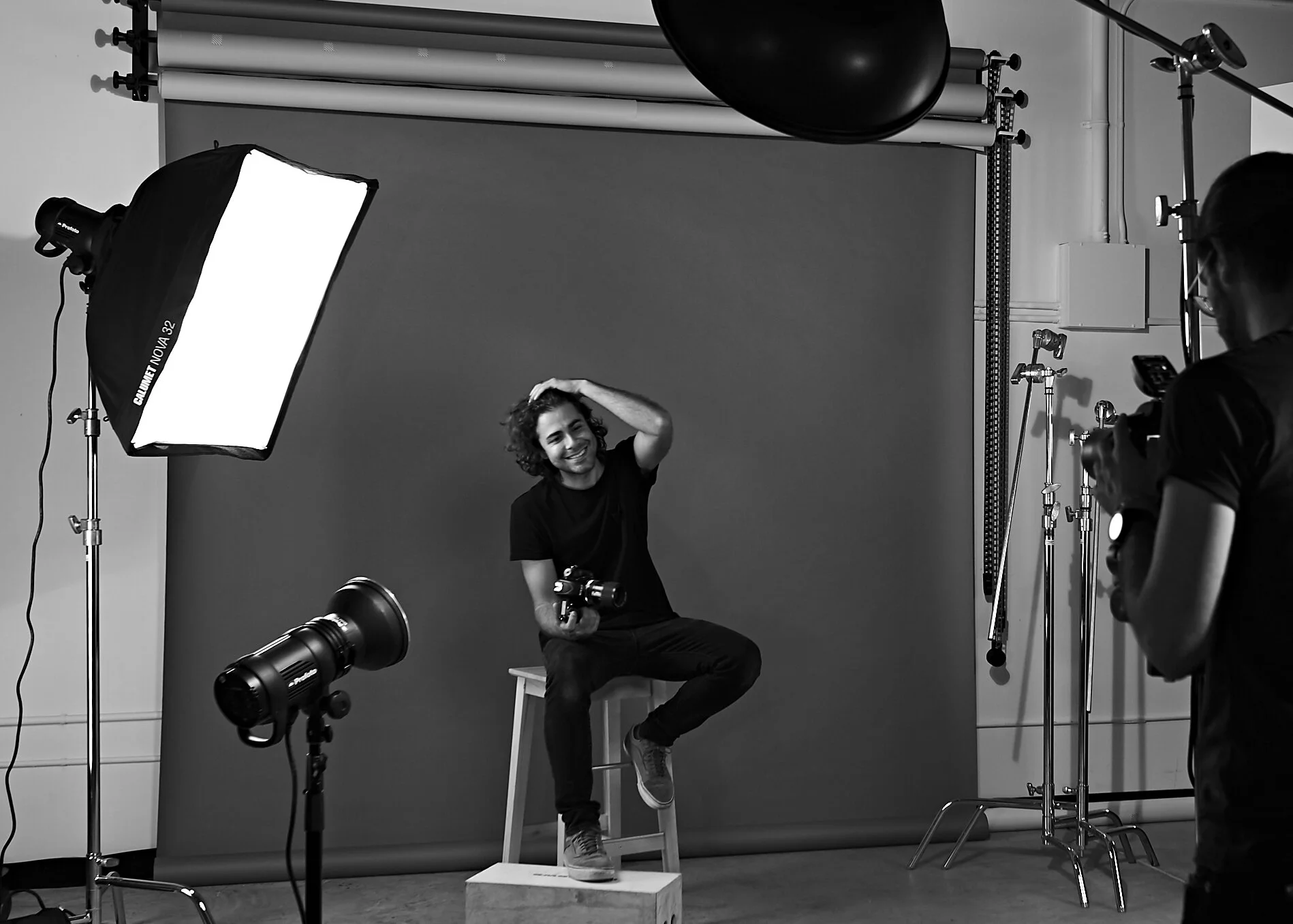 A man smiling and sitting on a stool in front of a photography backdrop during a photoshoot, with professional lighting and camera equipment around him.