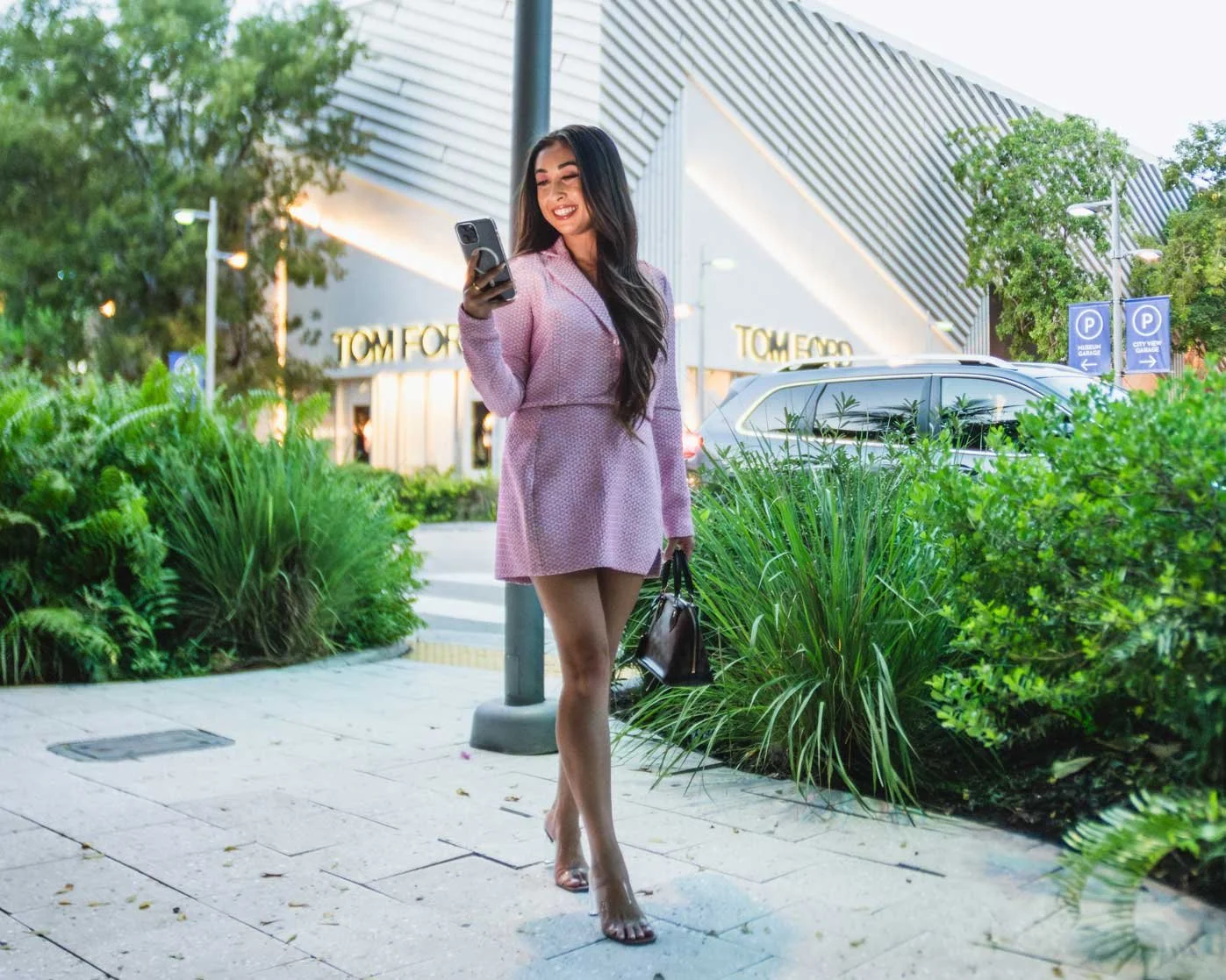 A woman in a pink dress walking on a sidewalk, looking at her phone, carrying a brown handbag, with a Tom Ford store and green landscaping in the background.