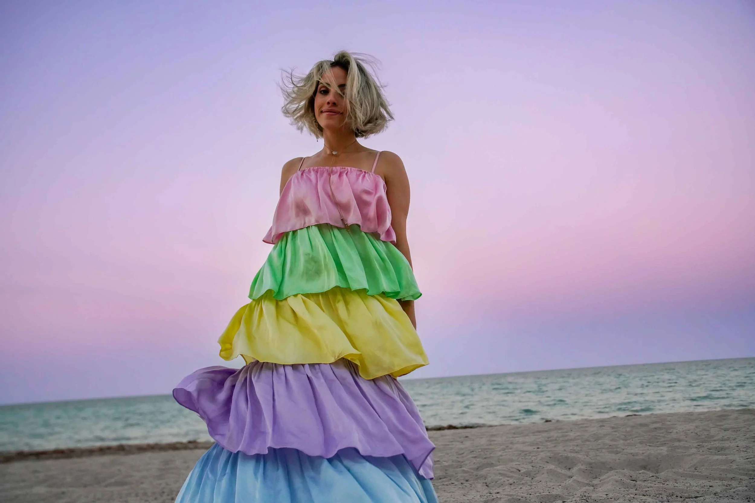 Woman on the beach wearing a colorful, tiered, pastel-colored dress with a pink, green, yellow, purple, and blue skirt, standing near the ocean with a pink and purple sunset sky in the background.