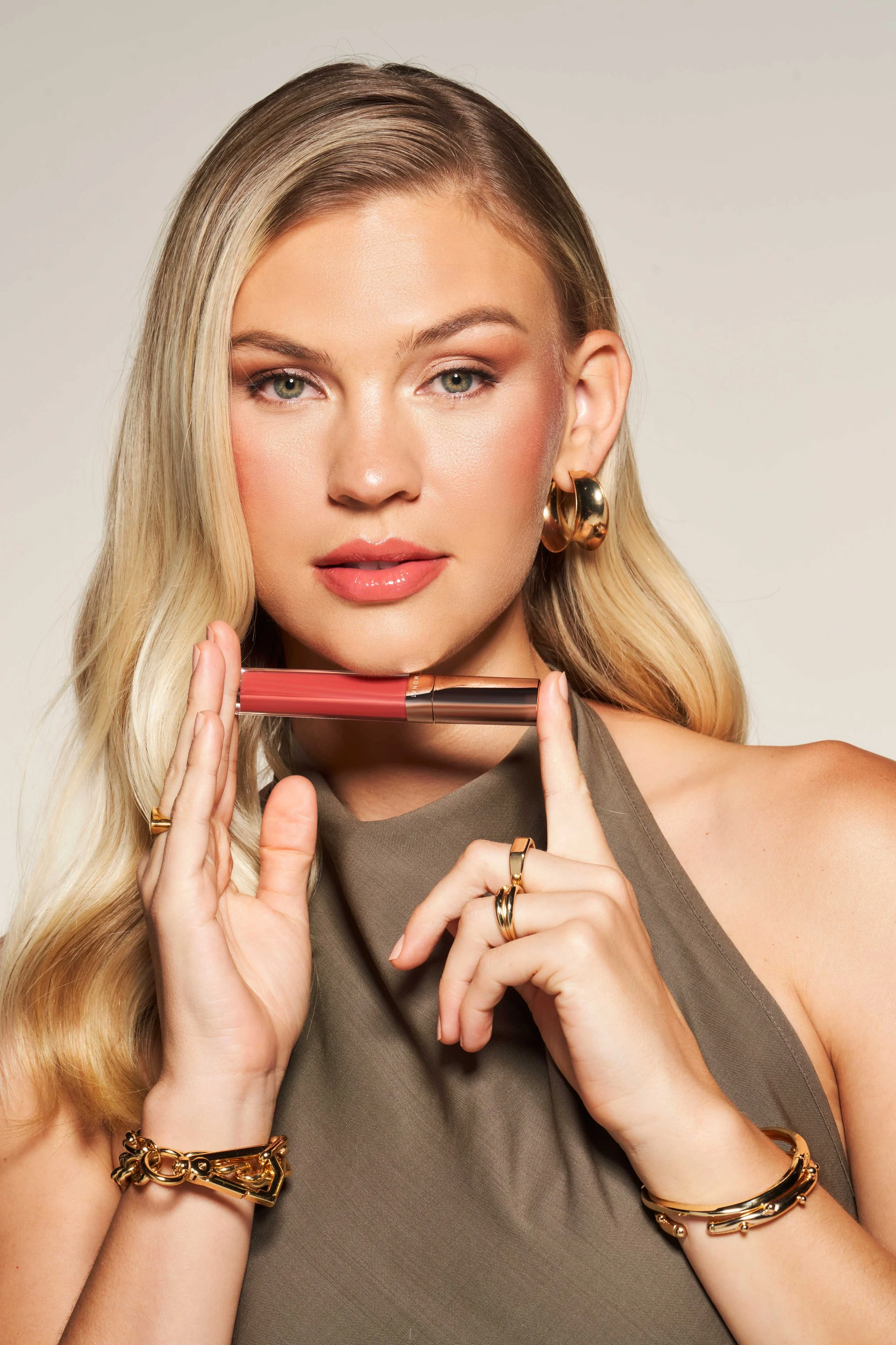A woman with blonde hair wearing gold jewelry and a sleeveless dress, holding a tube of lipstick.