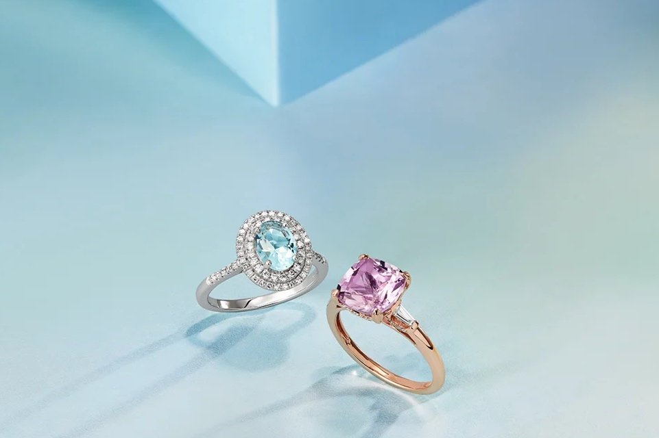 Two rings, one with a blue gemstone surrounded by smaller diamonds and the other with a pink gemstone, on a light blue and white surface.