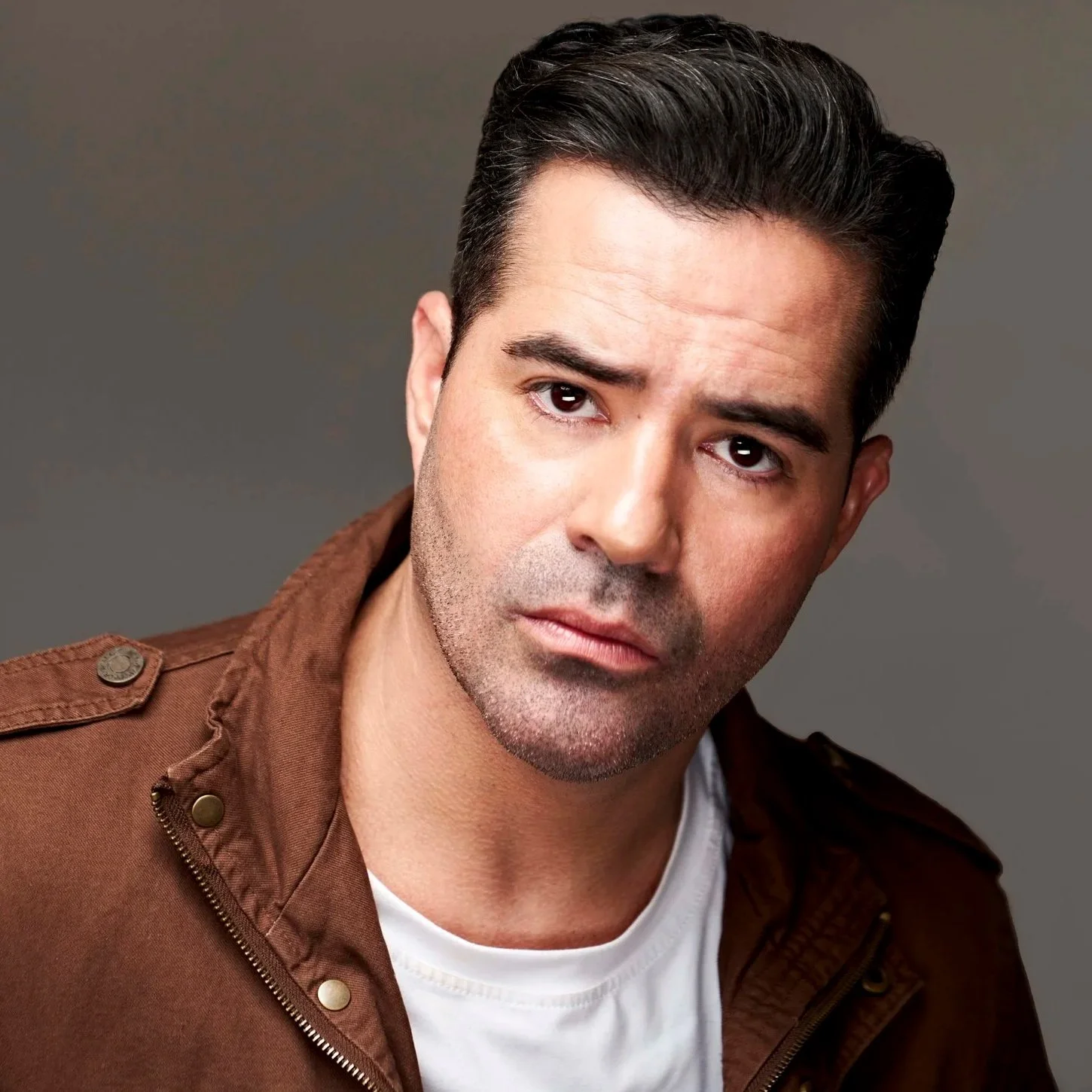 A man with dark hair wearing a brown jacket and white shirt against a neutral background.
