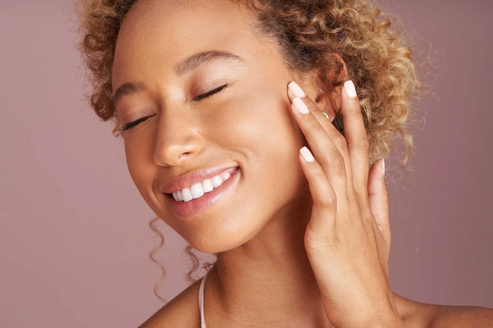 A woman with curly blonde hair is smiling with her eyes closed, touching her cheek with her hand, against a plain pink background.