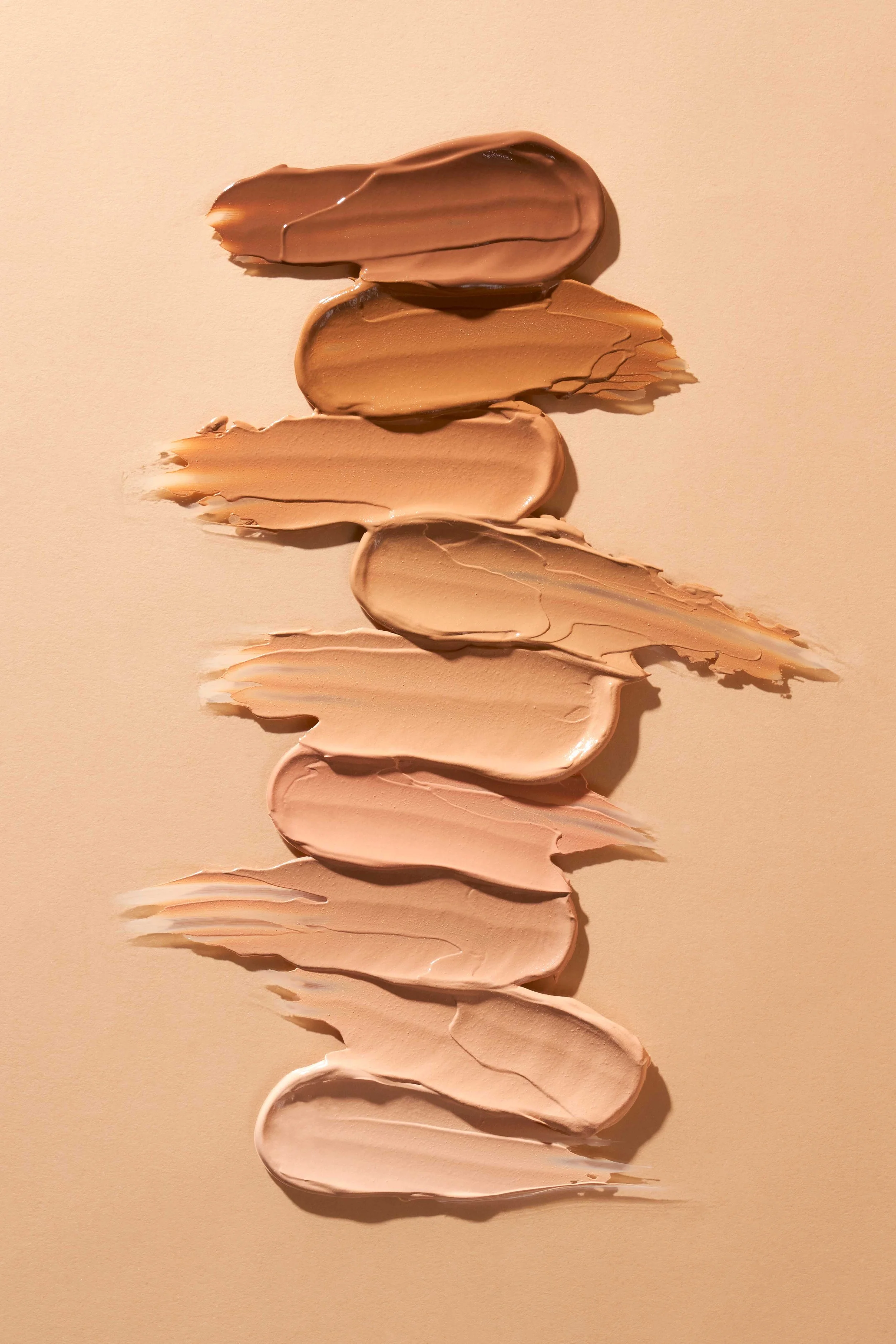 Swatches of various beige and brown foundation shades smeared on a beige background.