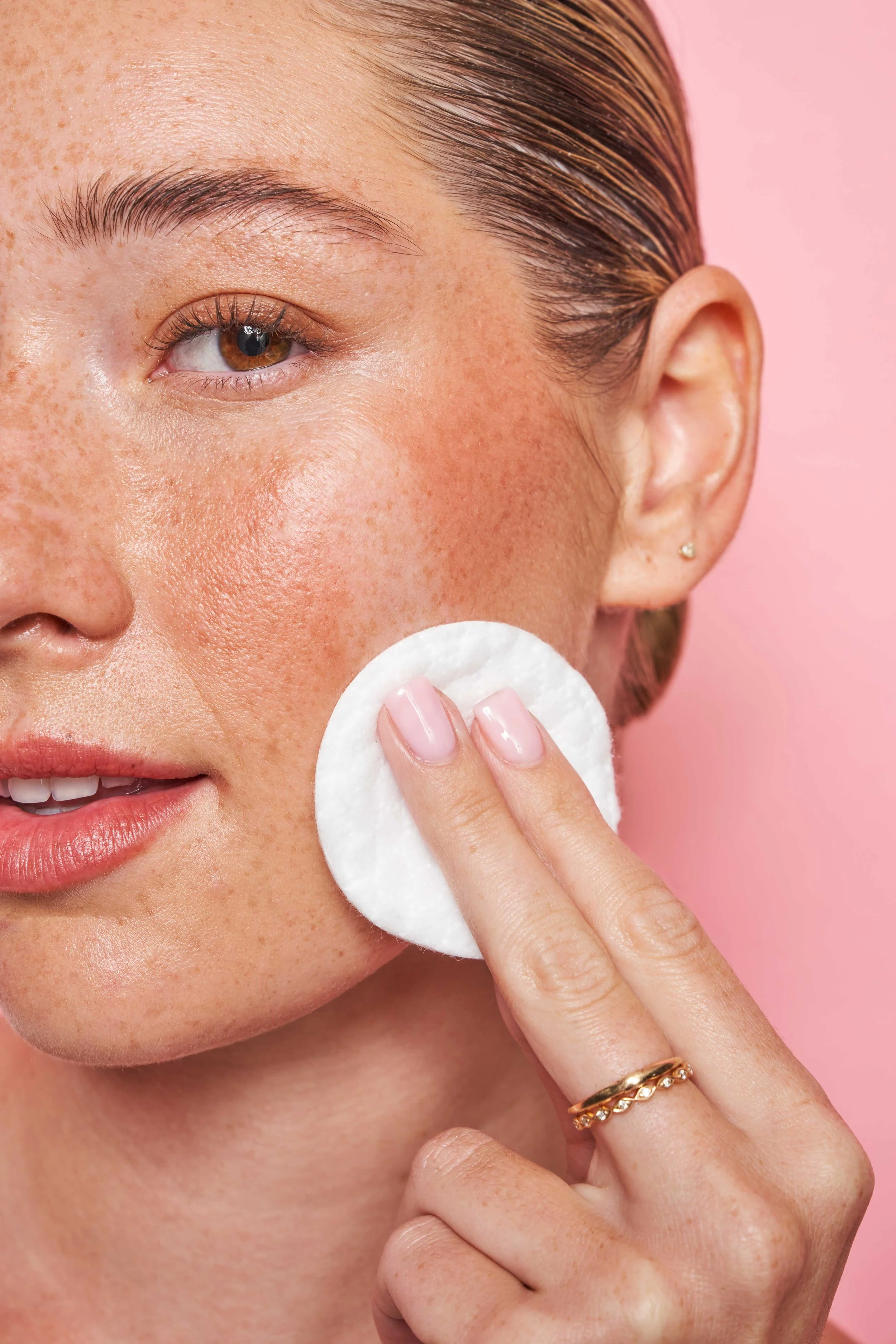 A close-up of a woman with light skin and freckles using a cotton pad on her cheek against a pink background.