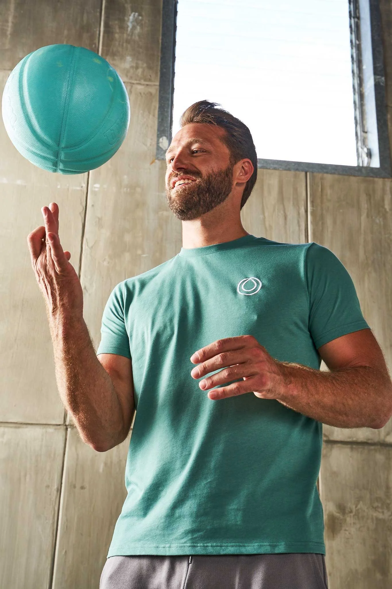 A man with a beard and short hair spinning a teal basketball on his finger indoors with a large open window behind him.