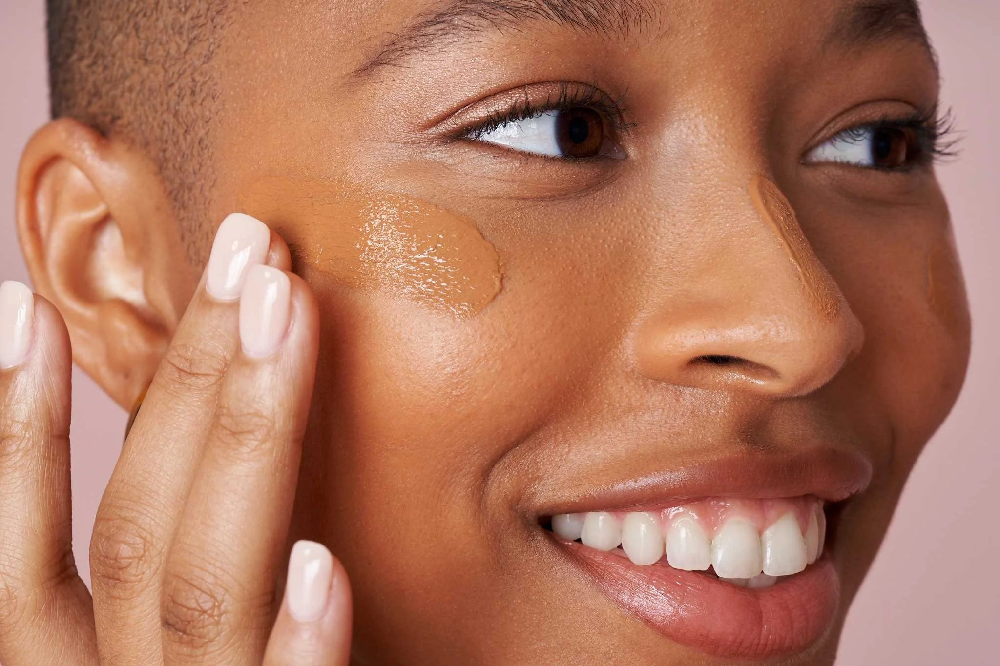 Close-up of woman applying foundation to cheek with finger, smiling, with warm-toned makeup and natural skin textured