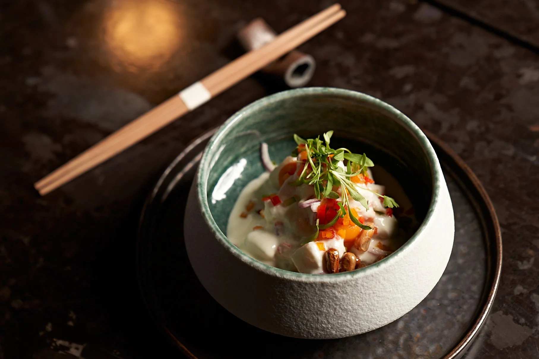 A bowl of Asian-inspired seafood salad with microgreens, chopped vegetables, and creamy dressing, placed on a dark table with chopsticks nearby.
