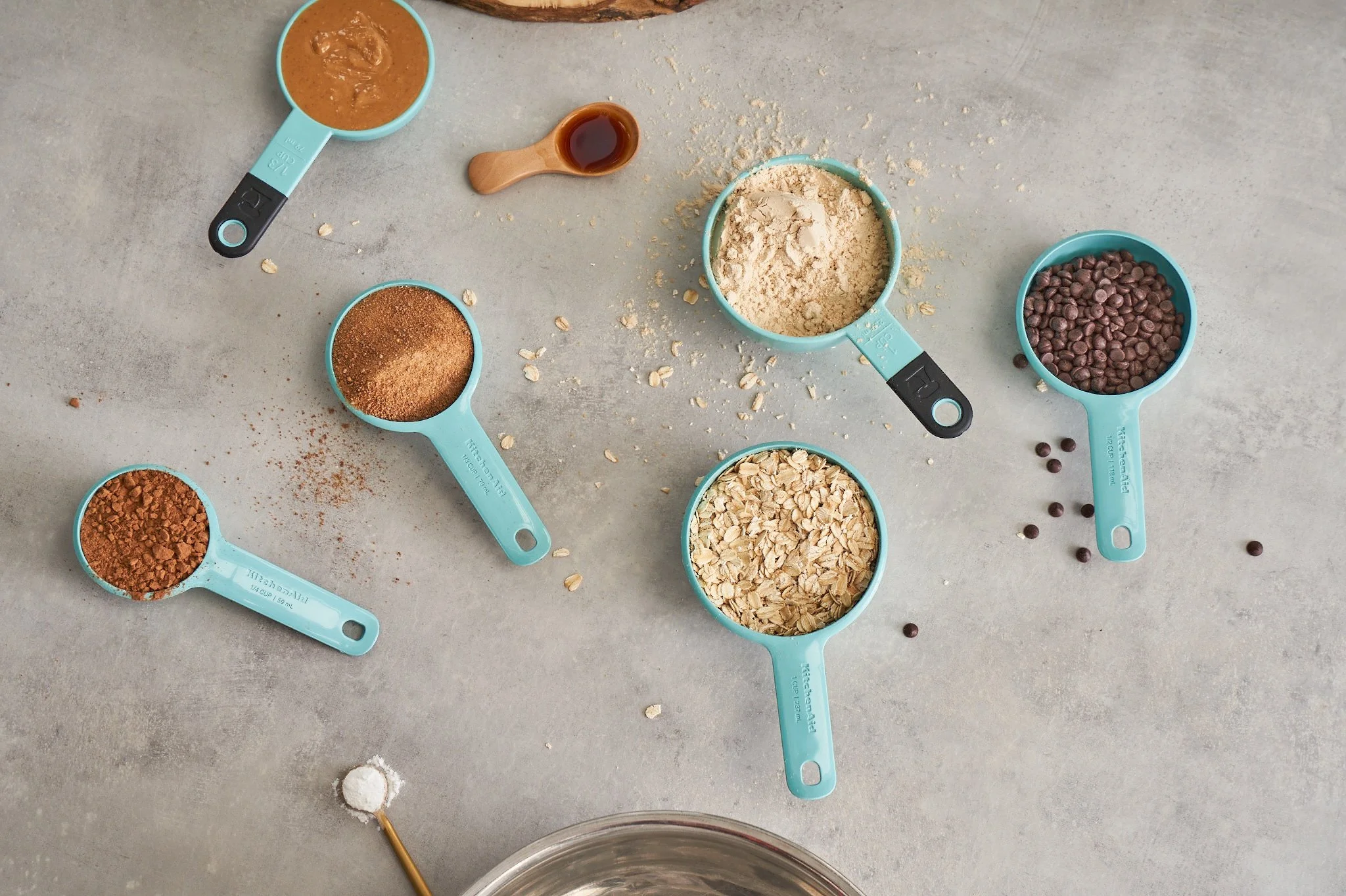 Various measuring cups filled with ingredients such as oats, chocolate chips, cocoa powder, and flavored powders, along with small scoops and a small wooden spoon, all arranged on a gray surface.