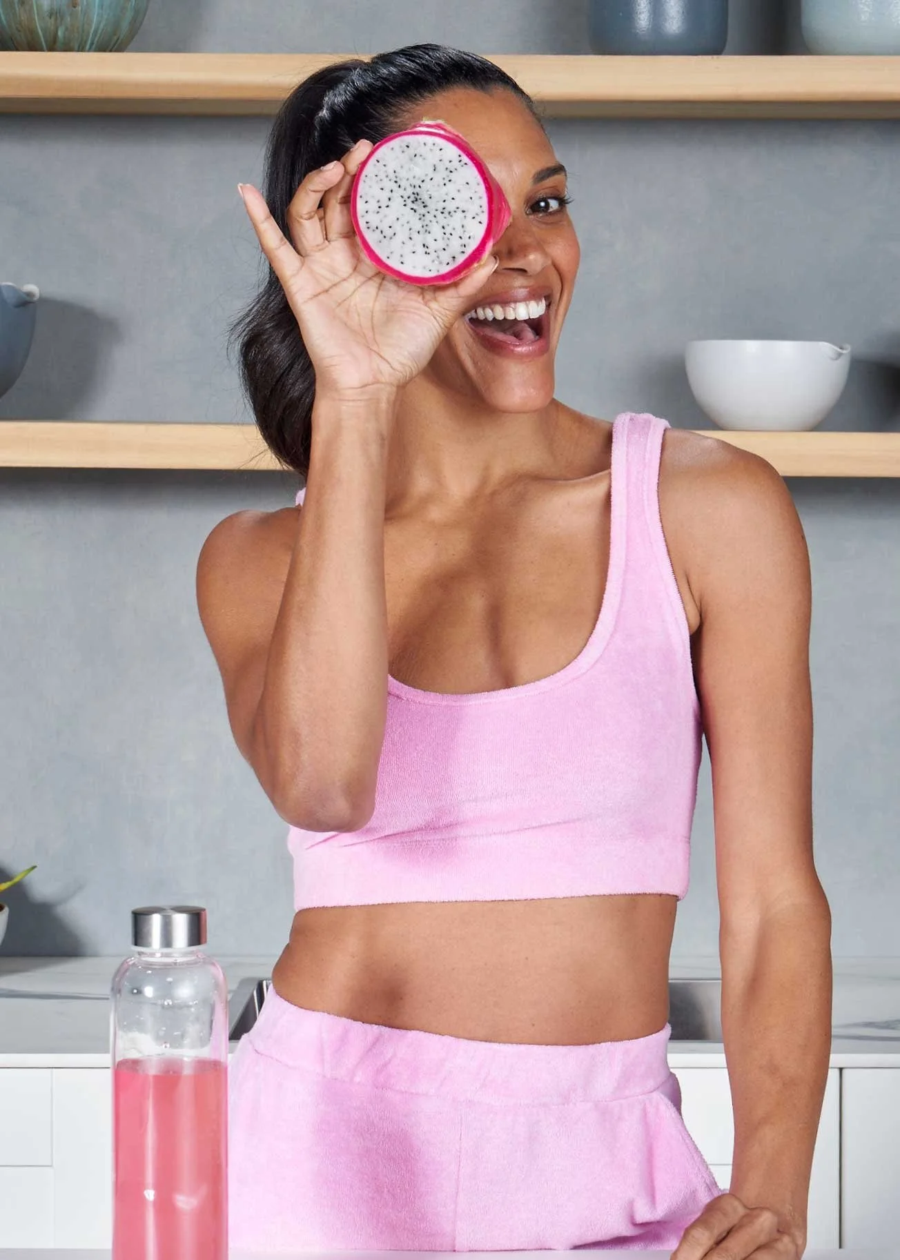 Smiling woman in pink sportswear holding a slice of dragon fruit over her eye in a modern kitchen.