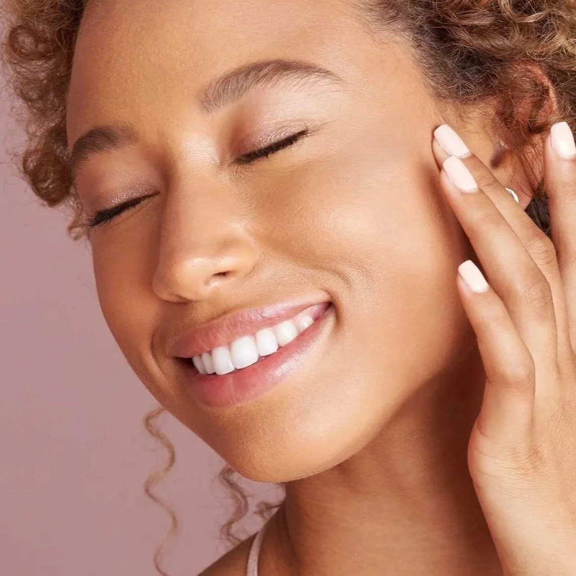 A close-up of a woman smiling with her eyes closed, touching her face with one hand. She has curly hair and is wearing light makeup.