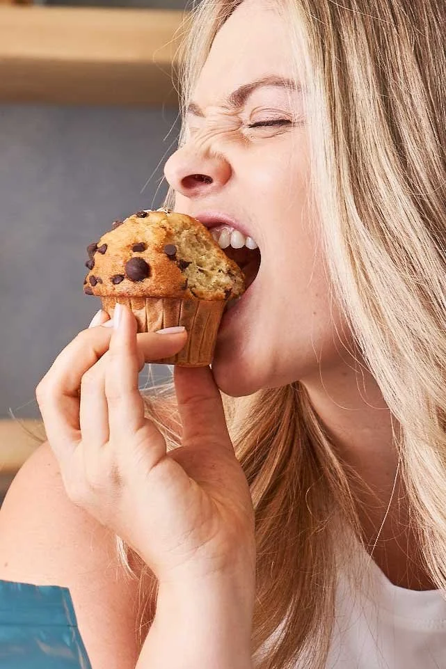 A woman with blonde hair is biting into a chocolate chip muffin.