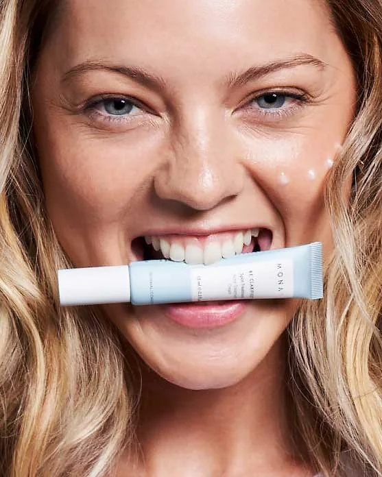 Close-up of a smiling woman with blonde hair holding a skincare cream tube in her mouth.