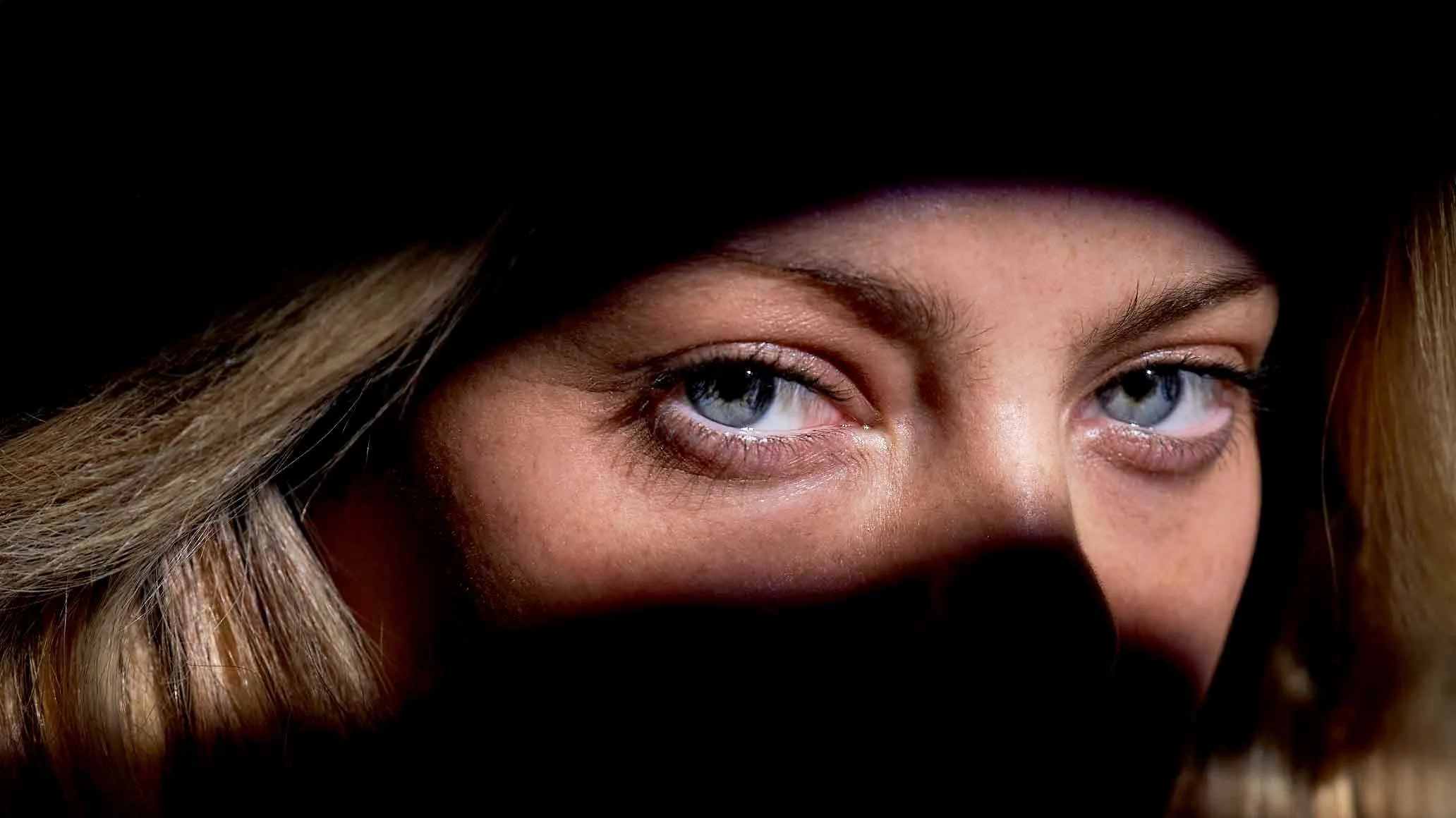 Close-up of a woman's face partially obscured by a black hat, revealing striking blue eyes and light brown hair.