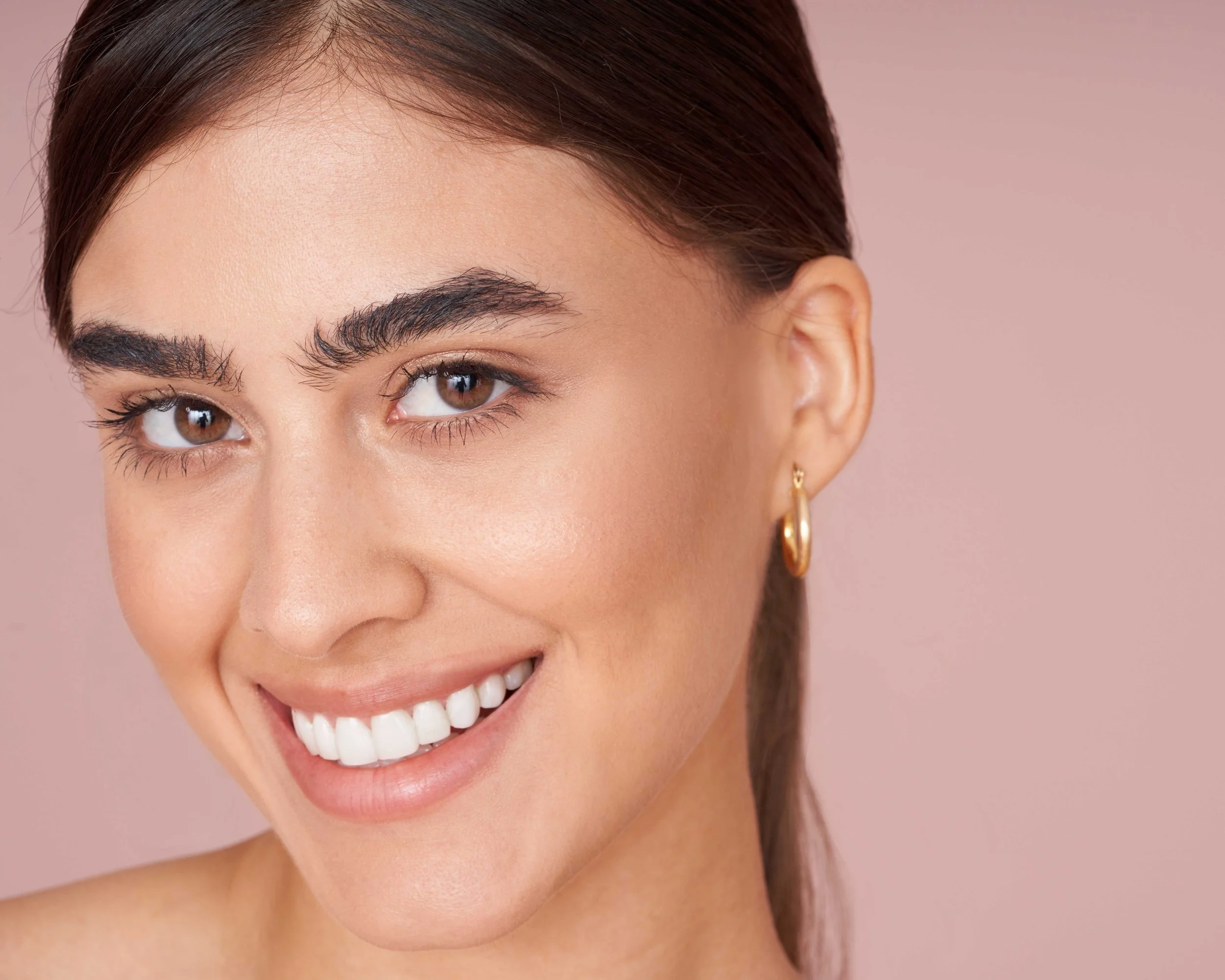 Close-up of a smiling woman with brown eyes, dark brown hair, gold hoop earrings, and light makeup against a pink background.