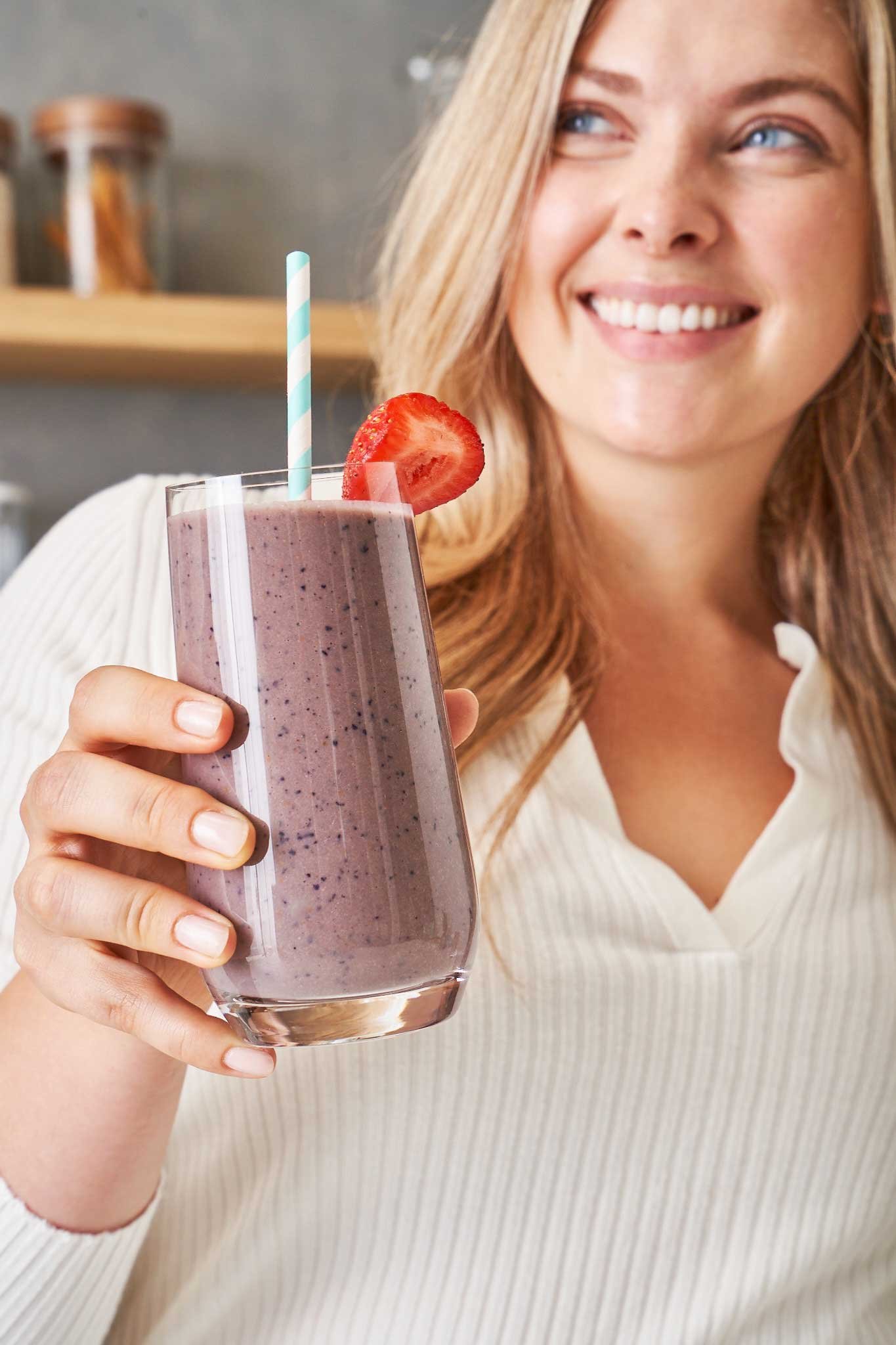 Woman holding a glass of purple smoothie with a strawberry slice and a blue-and-white striped straw.