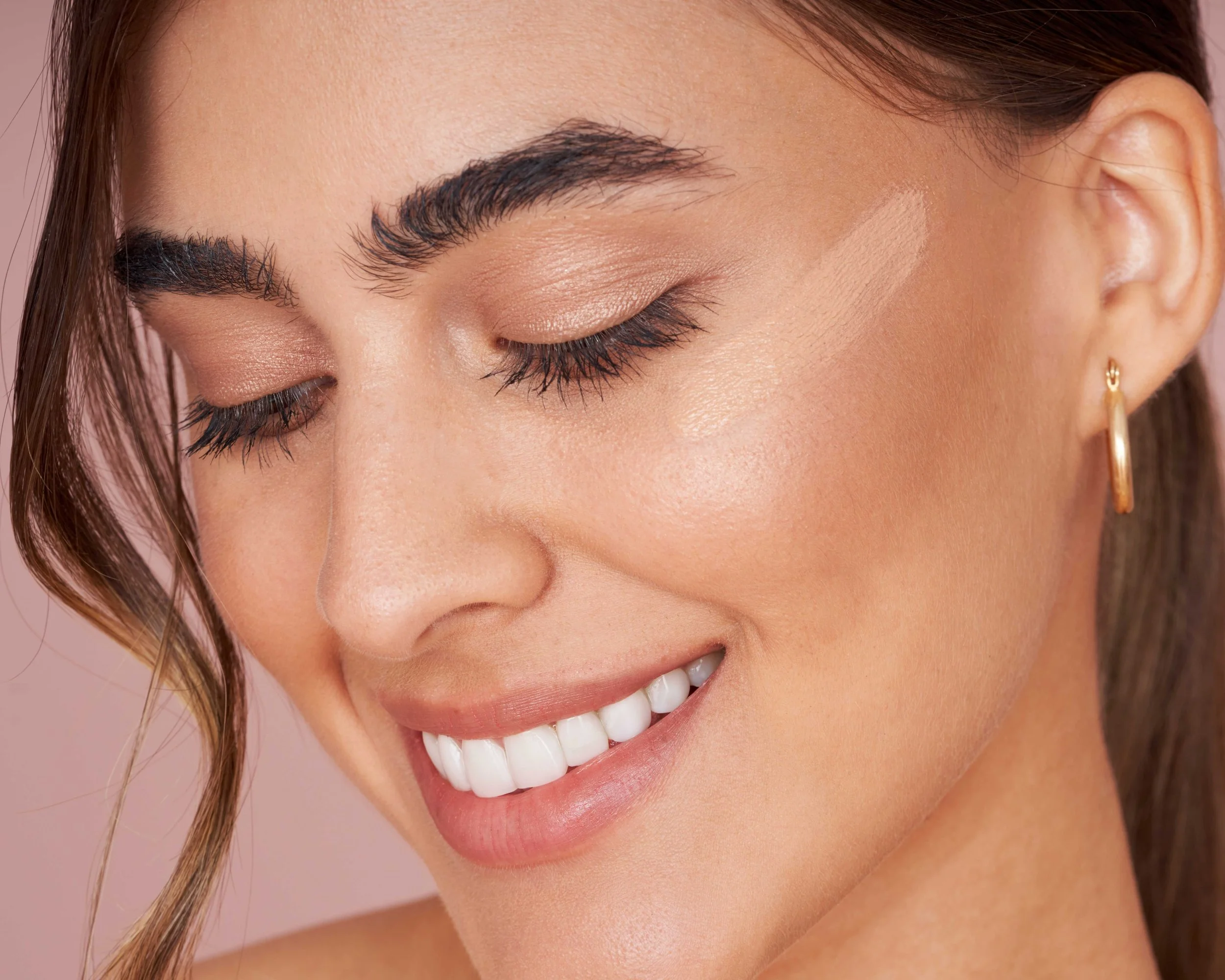 Close-up of a woman smiling with closed eyes, wearing natural makeup, gold hoop earrings, and light brown hair styled with loose waves.