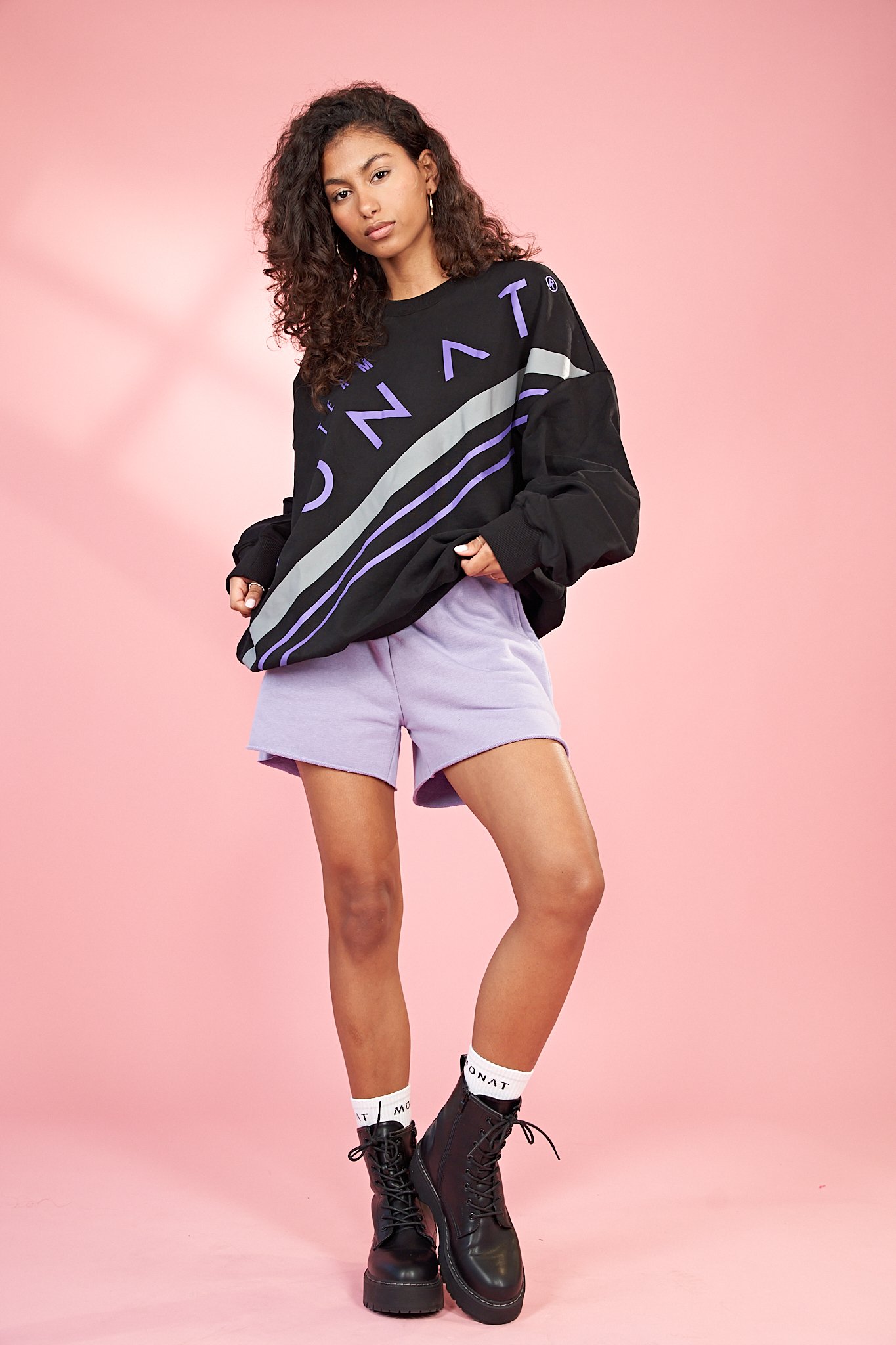 Young woman with curly hair wearing a black graphic sweatshirt, lilac shorts, white socks, and black combat boots, standing against a pink background.