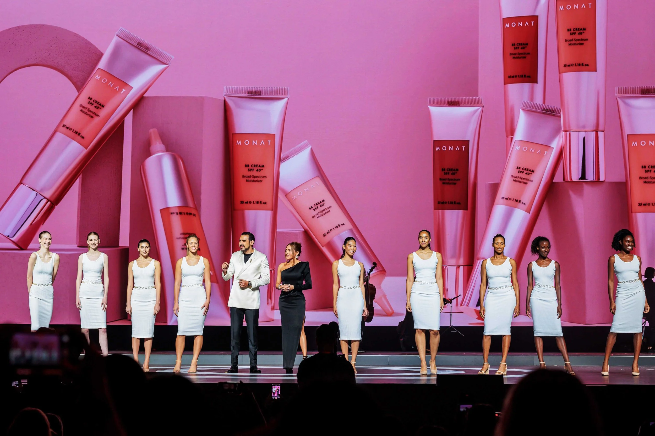 A group of models and presenters on stage at a fashion or beauty event, with a large pink background displaying oversized skincare product images labeled 'MONAT' and 'Broad Spectrum Moisturizer' with SPF 40.