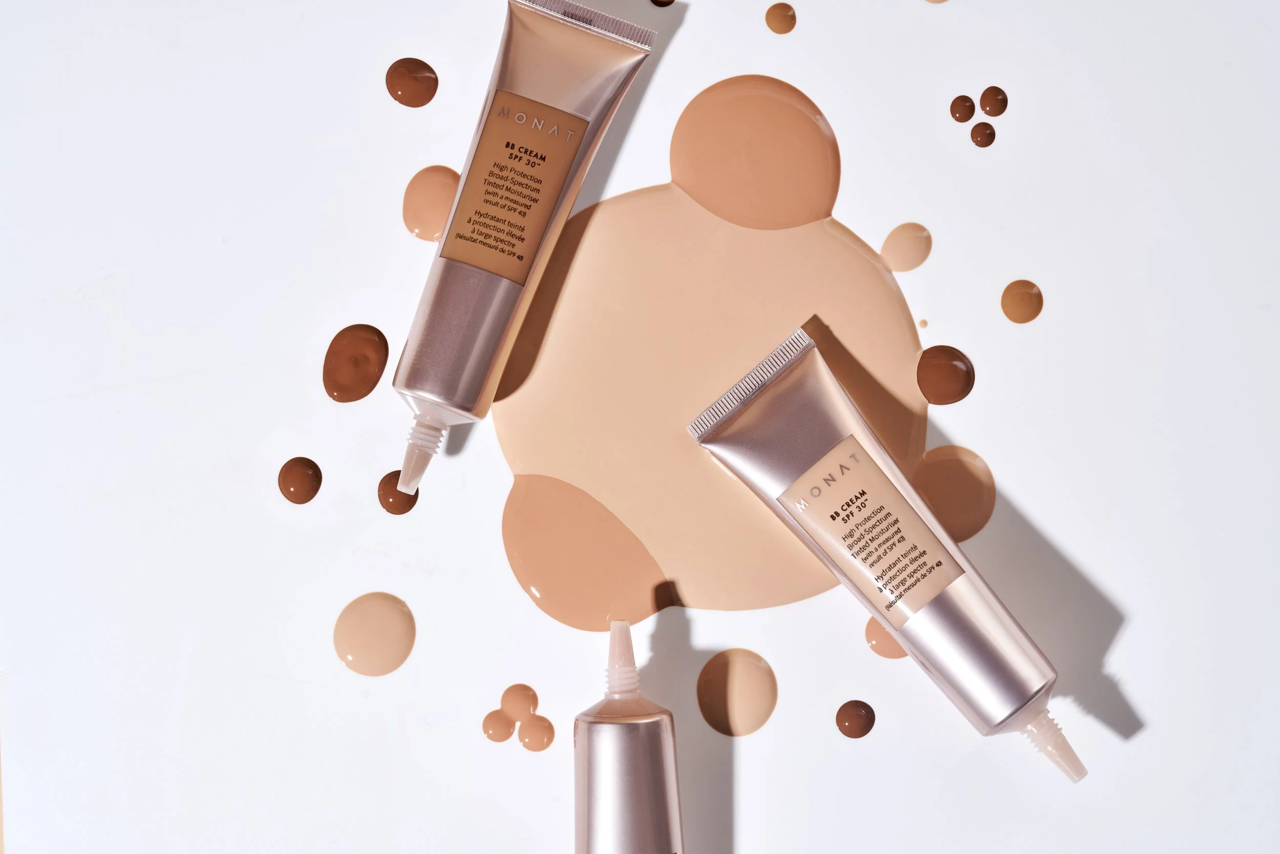 Two tubes of Monet BB cream with SPF 30 lying on a white surface surrounded by various drops and blobs of different shades of beige and brown foundation makeup.