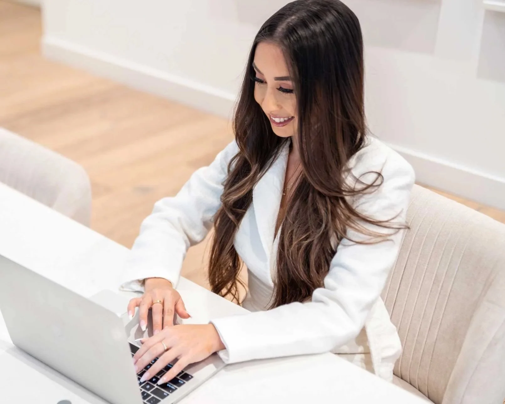 A woman with long, wavy brown hair, wearing a white blazer, sitting at a white desk, working happily on a silver laptop in a bright, modern room.