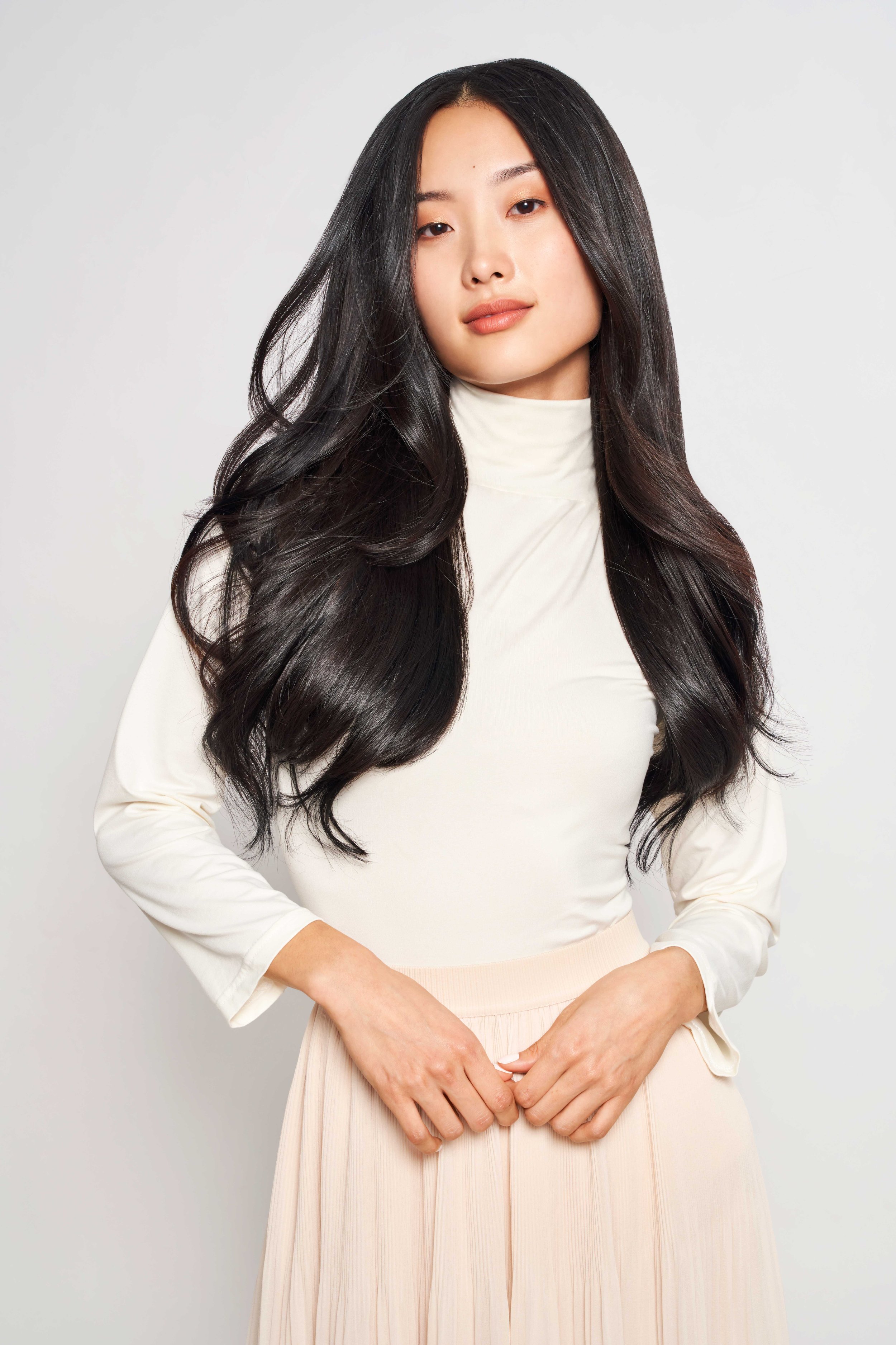 A woman with long, wavy black hair and light makeup, wearing a high-neck cream top and a light-colored skirt, standing against a plain light background.