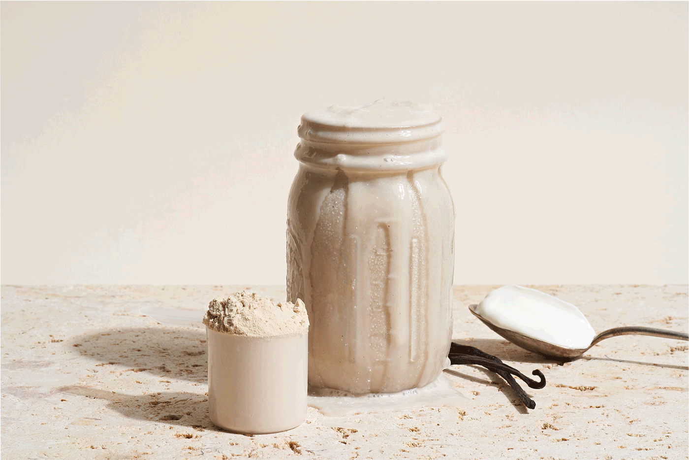 A jar of vanilla protein powder with a scoop of powder and vanilla beans on a light-colored surface