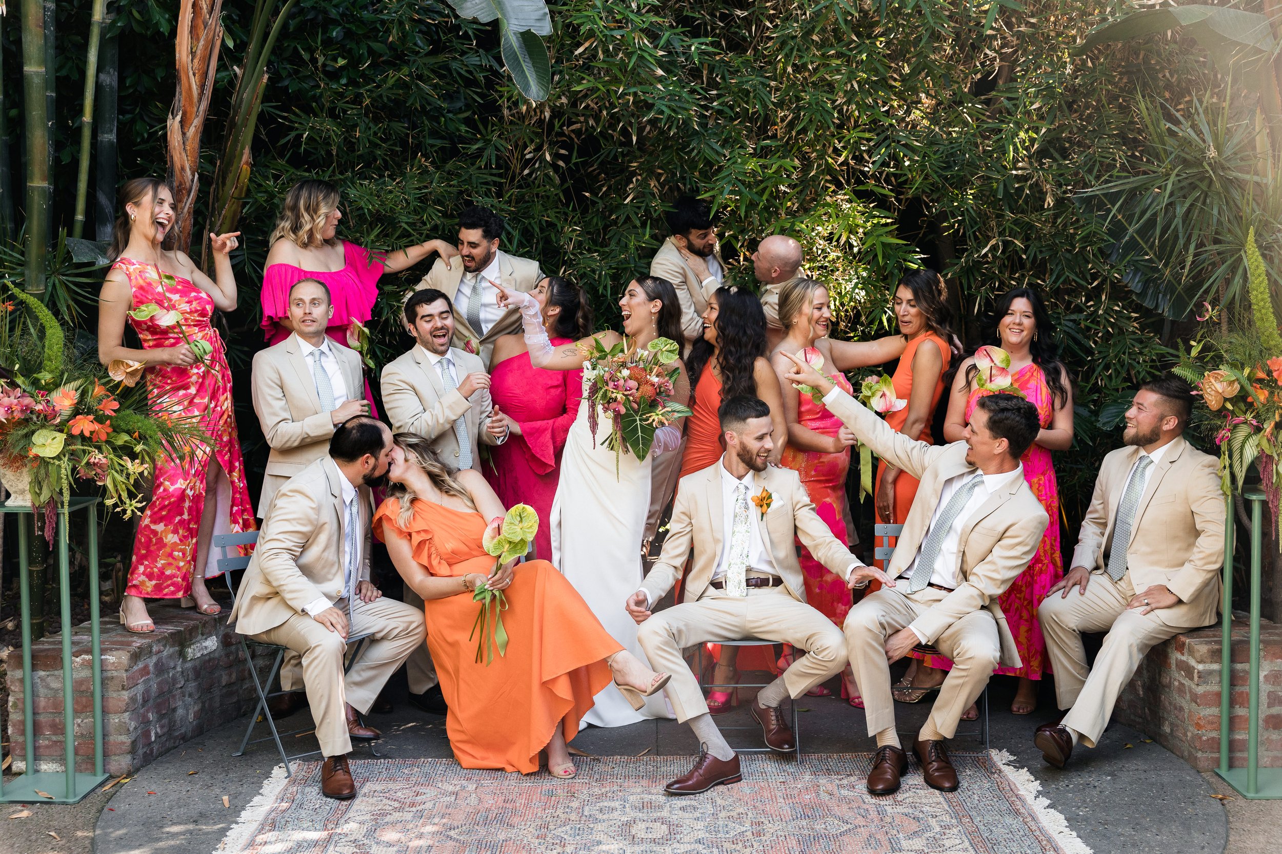 A group of wedding guests wearing colorful dresses and suits gathered outdoors among lush green plants, celebrating and dancing.