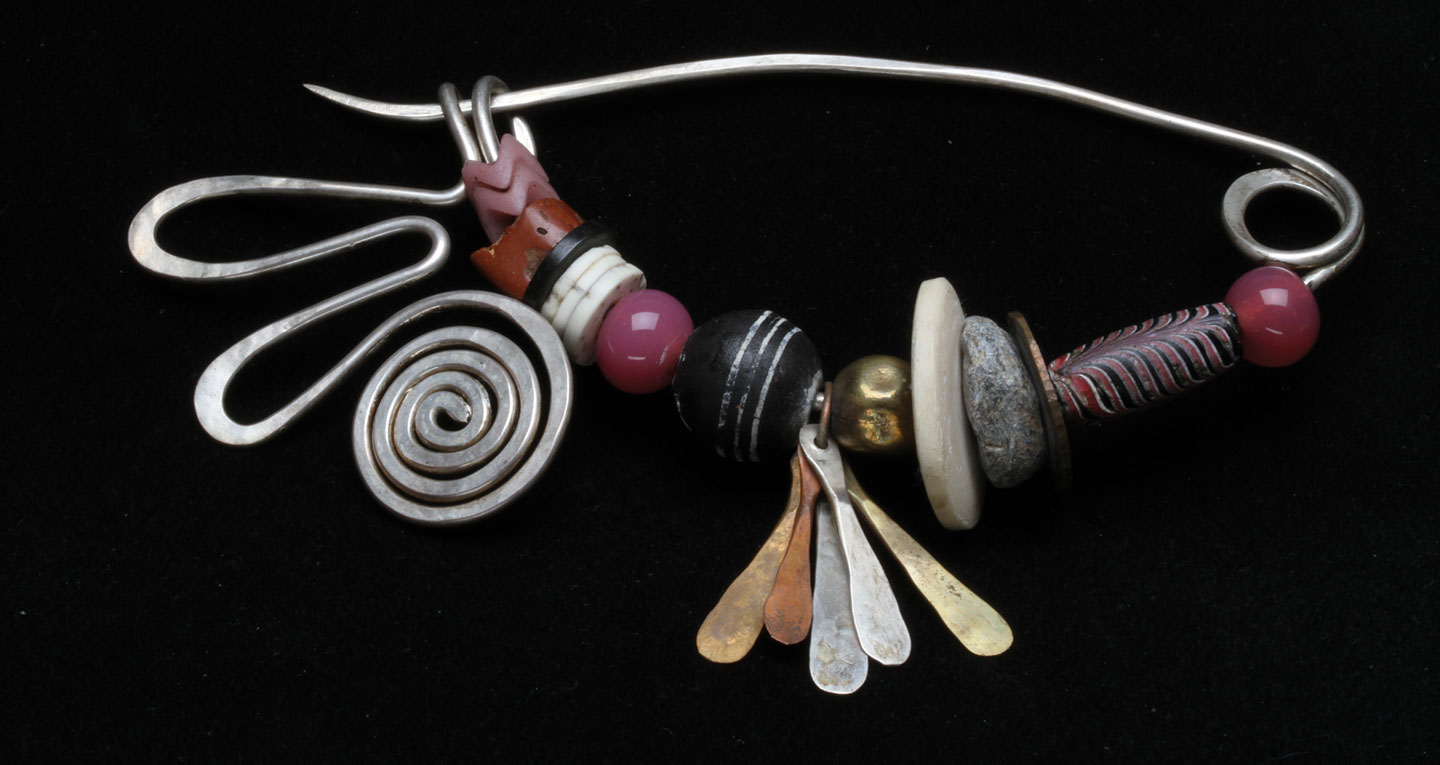 FIBULA PIN of sterling silver wire, formed, forged and filed, strung with molded Czech glass snake beads, bauxite bead, coconut shell disk bead, ostrich shell disk beads, vintage Chinese glass beads, African terracotta spindle whorl; forged/drilled dangles of silver, bronze and copper, metal bead, stone beads, copper metal disk, Venetian glass bead, many from the African trade; 13.6 centimeters wide, dating from mid-1980s. Spring portion of pin forged to work harden it; priced eighty-five dollars when purchased in 1988. Carolyn L. E. Benesh/Ornament Collection. Photographs by Robert K. Liu/Ornament.