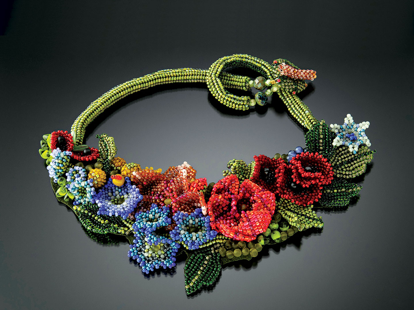 BLUEBELLS AND POPPIES FIORI NECKLACE of glass beads, woven in various off-loom stitches with a needle and fishing line in a free-form style. Petals and leaves are woven and then embroidered to each other and a fabric base. Embellished with stones and fire-polished glass, the necklace is backed with Ultra-suede; back of necklace is created with a filled herringbone spiral tube, 43 centimeters long x 9 centimeters high, 2014.