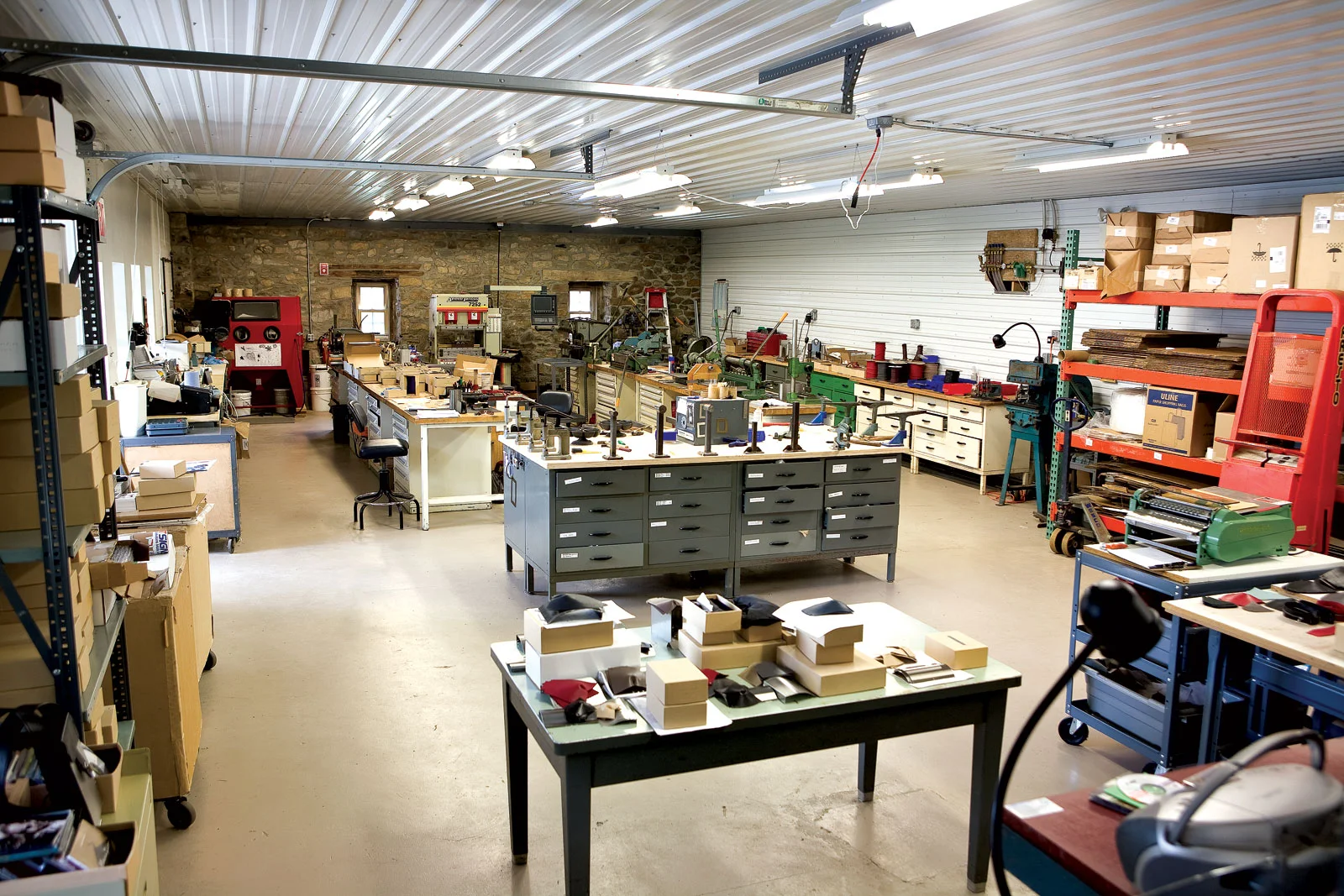 WENDY STEVENS STUDIO. After the studio fire in 2004, the facility was redesigned and rebuilt on the main level of the stone barn on her eight-acre farm property. In the foreground is a packing table with finished pieces. To the right is the leather cutting table with a gluing machine. Directly behind the packing table is the stake bench where the pieces are assembled. Against the wall are the orange pallet racks where sheet metal is stored with a hydraulic lift. In the background are the tool benches with slip rolls, notchers, shears, a hand press brake and a hydraulic brake, a sand blast cabinet and deburring wheels. Photograph by Kate Lacey