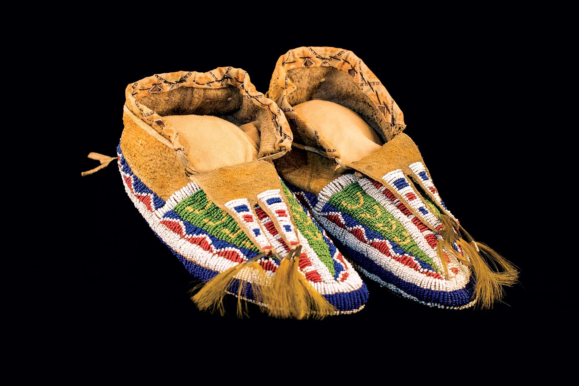MOCCASINS WITH BEADED SOLES (Sioux) of cowhide, glass beads, sinew, tin tinklers, cow tail hair, prior to 1890. Commonly thought to be for use in burials, moccasins with beaded soles were in actuality a way to honor living people. They were used in ceremonies, to recognize individual achievement and to show status. Some have wear on the soles, confirming that they were worn to walk on.