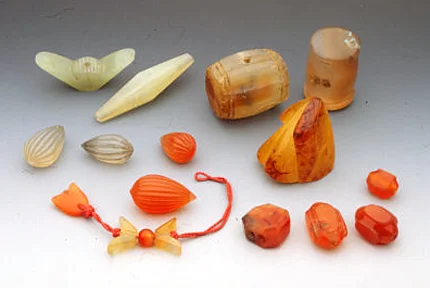 Chinese jade imitations and carnelian beads and ornaments in traditional shapes. One of the jade imitation is in the shape of Chinese money, at least three are toggles used by men, and the others are probably used for dangles, including two shaped as money. At least four are paneled carnelian beads. RKL