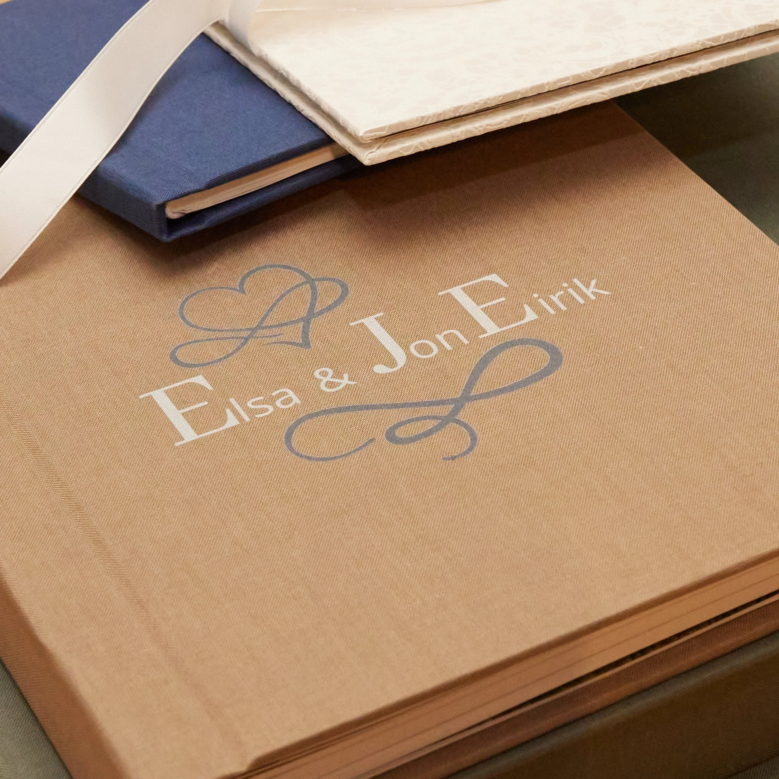 Ett bryllupsalbum med texten 'Elsa & Jon Erik' og dekorative elementer, samt noen andre album på en bord.