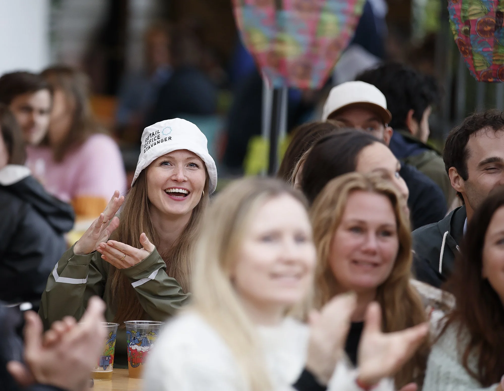 En gruppe folk smiler og ler, mens de sitter på en samling, noen med klare ansikter og andre dillet. En kvinne med lang blond hår og hvit hatt som har teksten 'Retail Office Stavanger' er i fokus.