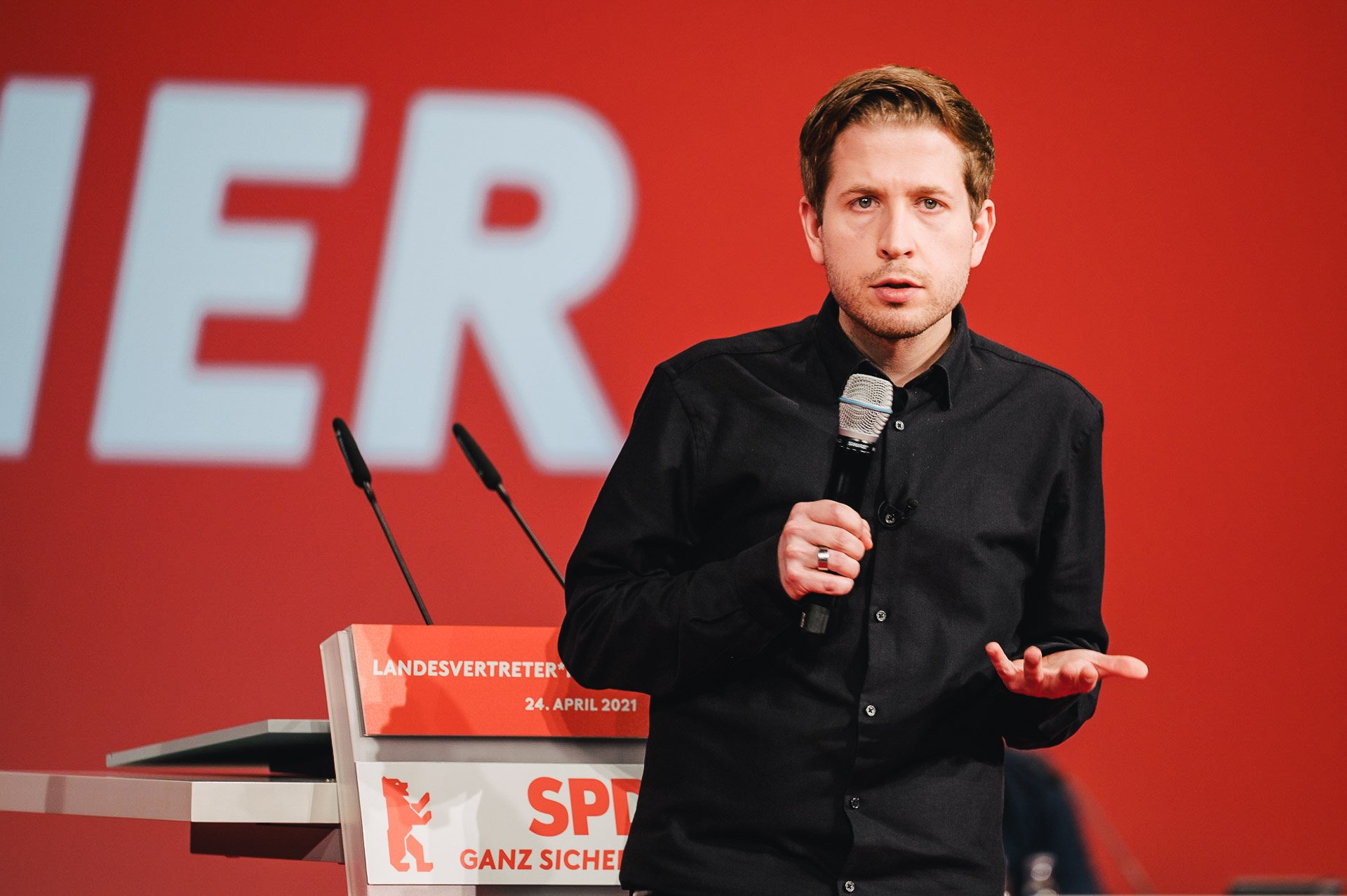  Kevin Kühnert spricht auf dem Landes Parteitag der SPD Berlin. Er hält ein Mikrofon in der Hand und gestikuliert. Vor einem roten Hintergrund. 