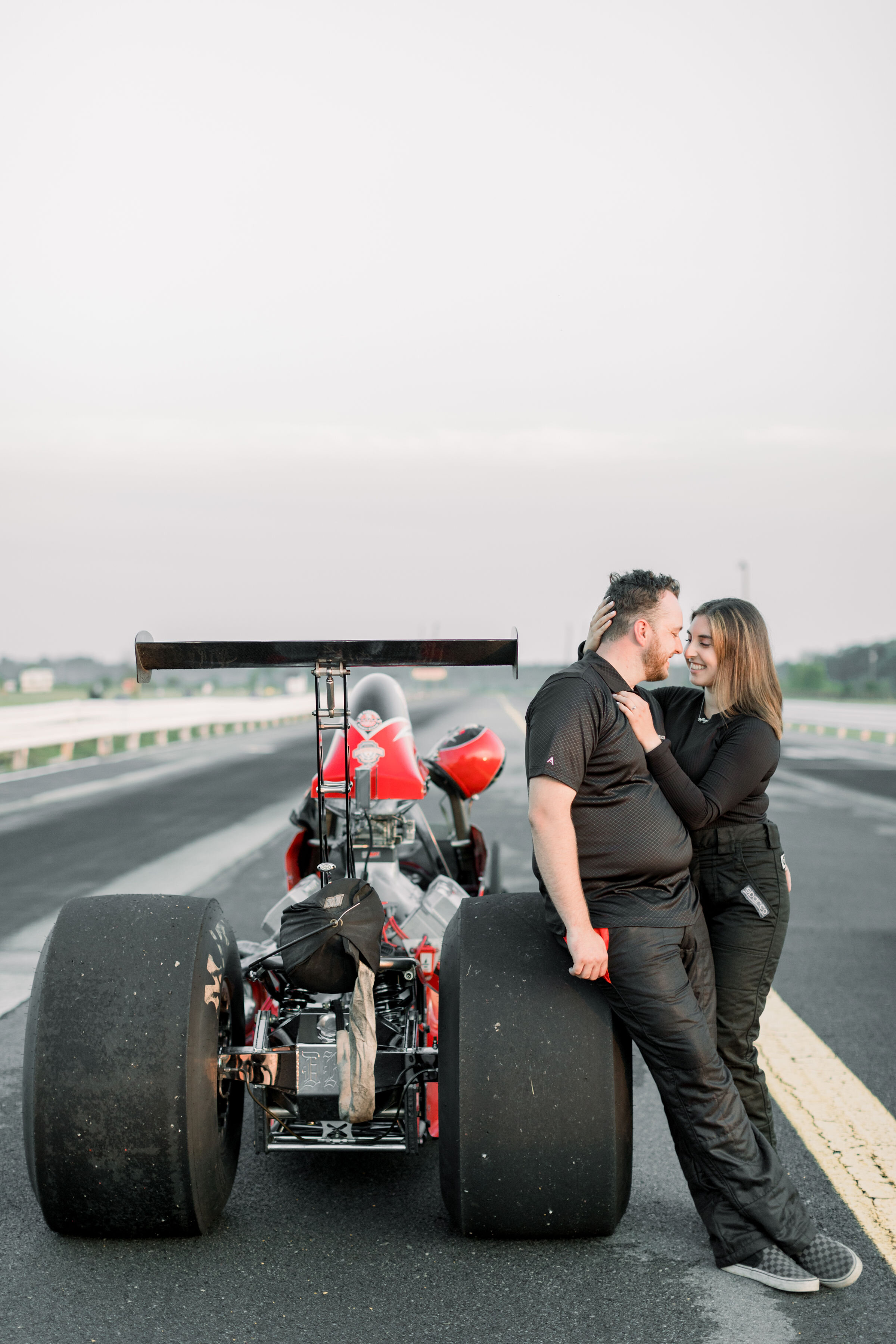  Happy couple leaning on car that is on a racetrack for engagement photos by Chelsea Mason in Ottawa, ON.  engagement poses with cars unique engagement photos how to use props in photos how to pose couples in engagements best photographer in ottawa c