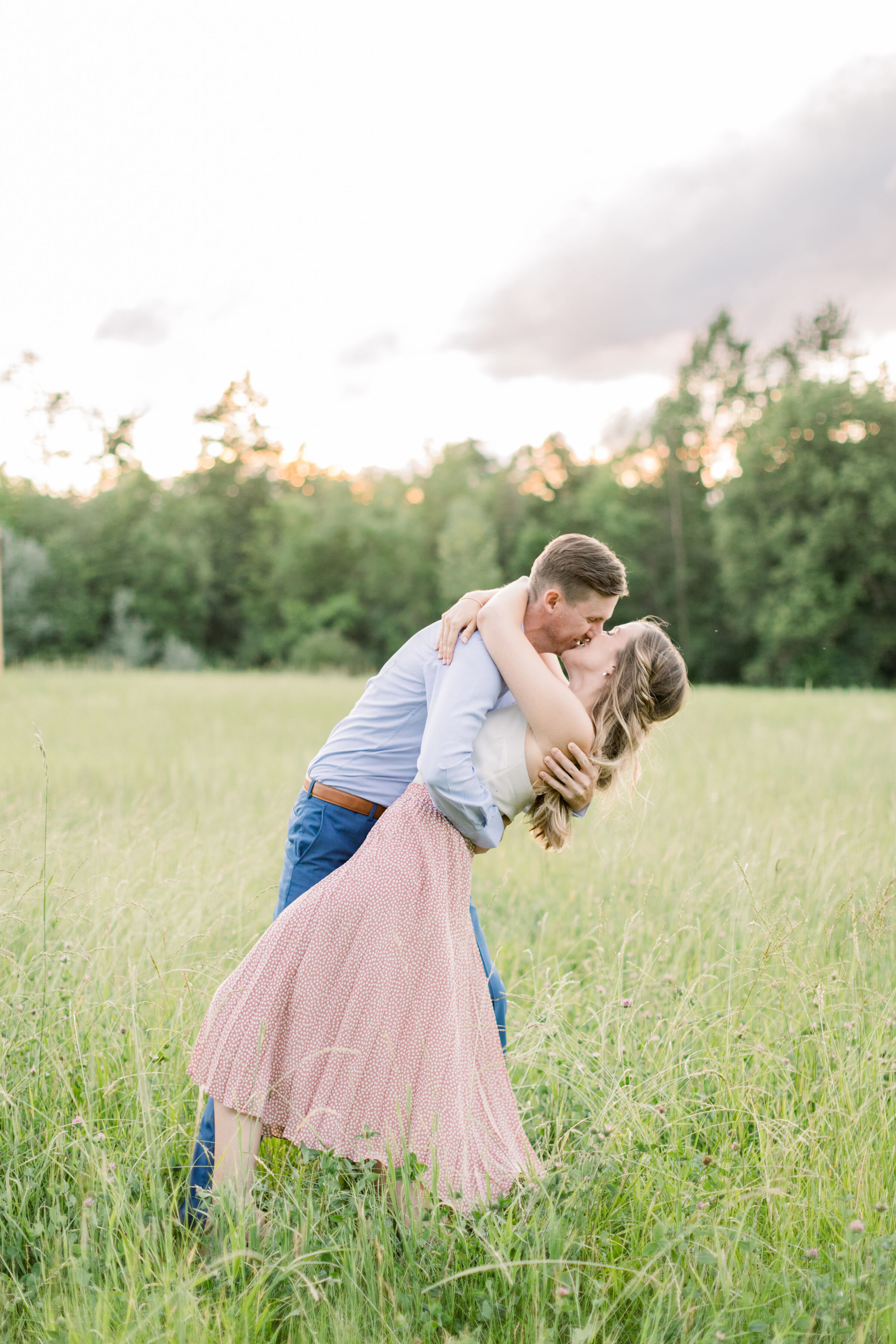  Couple doing a dip kiss pose wearing white and pink and blue in a green field during a summer engagement photoshoot in Ottawa, ON by Chelsea Mason Photography. kissing couple poses cute engagement poses for engagements outdoor locations for photosho