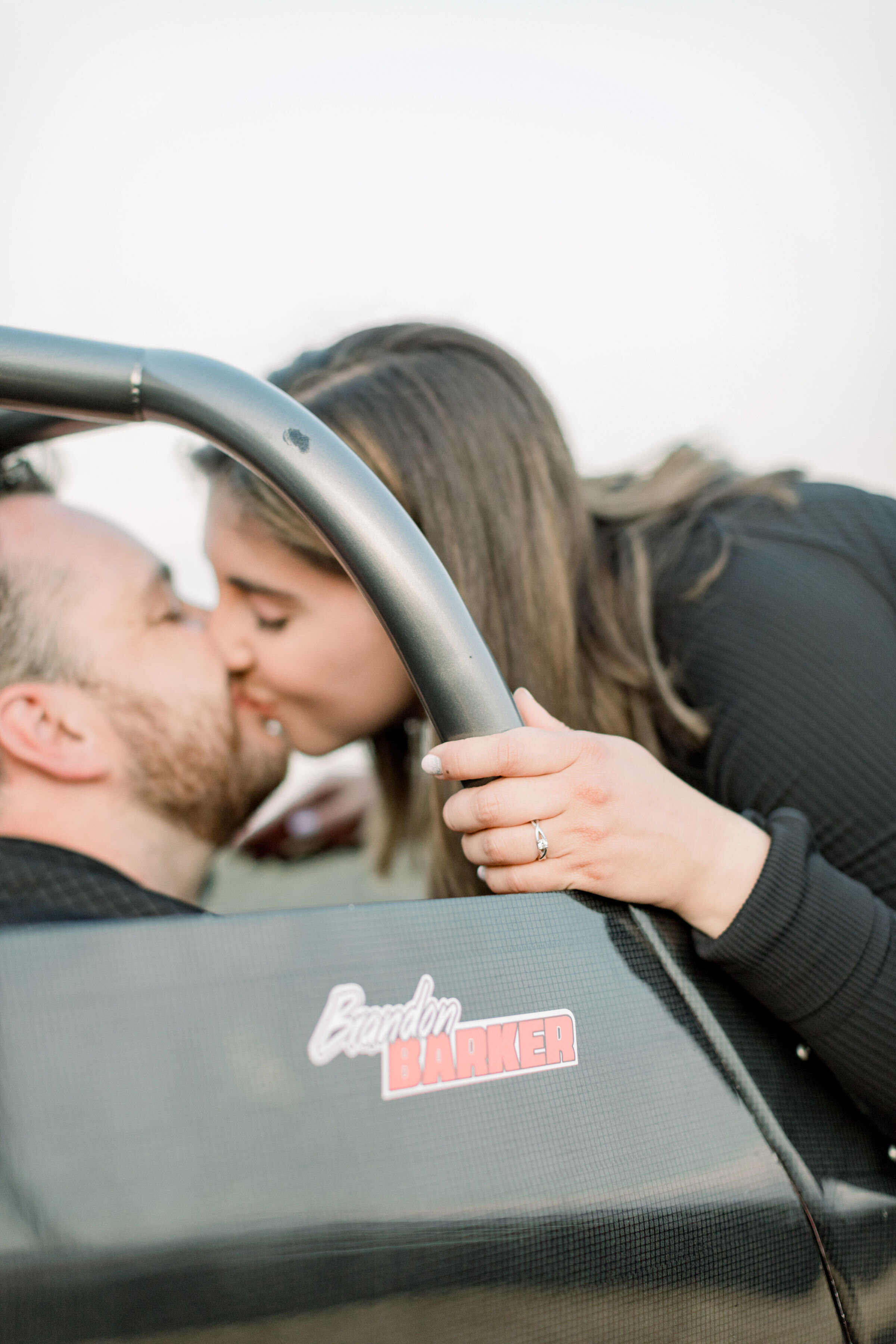  Detail shot of engagement diamond ring with a couple kissing in a car at a racetrack by Chelsea Mason in Ottawa, ON. wedding ring ideas engagement ring inspo simple wedding rings for bride wedding ring photography detail shot car in engagement photo