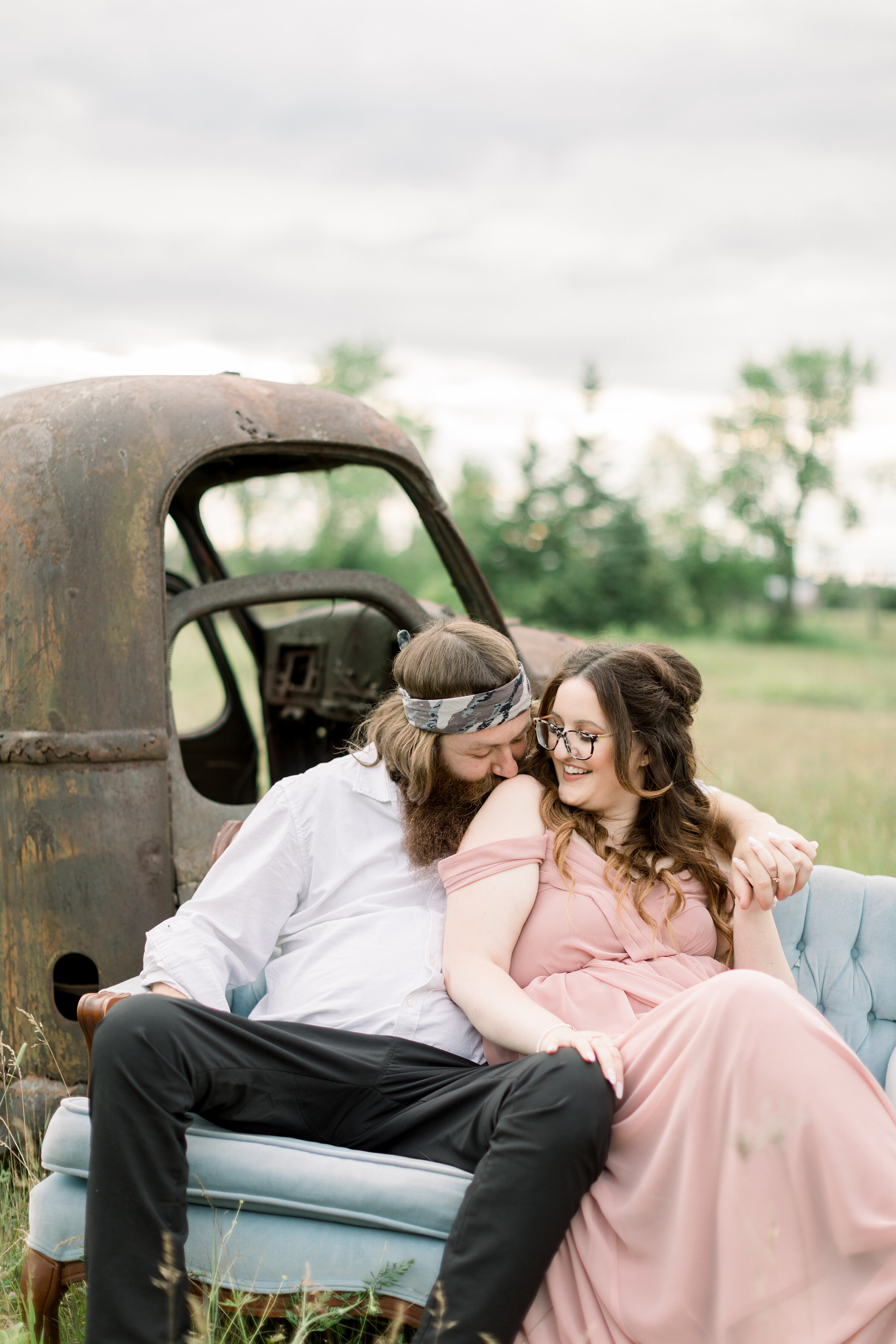  A loving couple sit together on a blue tufted couch by a rusting old truck in an awesome engagement session by Chelsea Mason Photography in Smith Falls, Ontario Canada. Sitting couple pose inspiration photo shoot prop goals outdoor photo shoot inspi