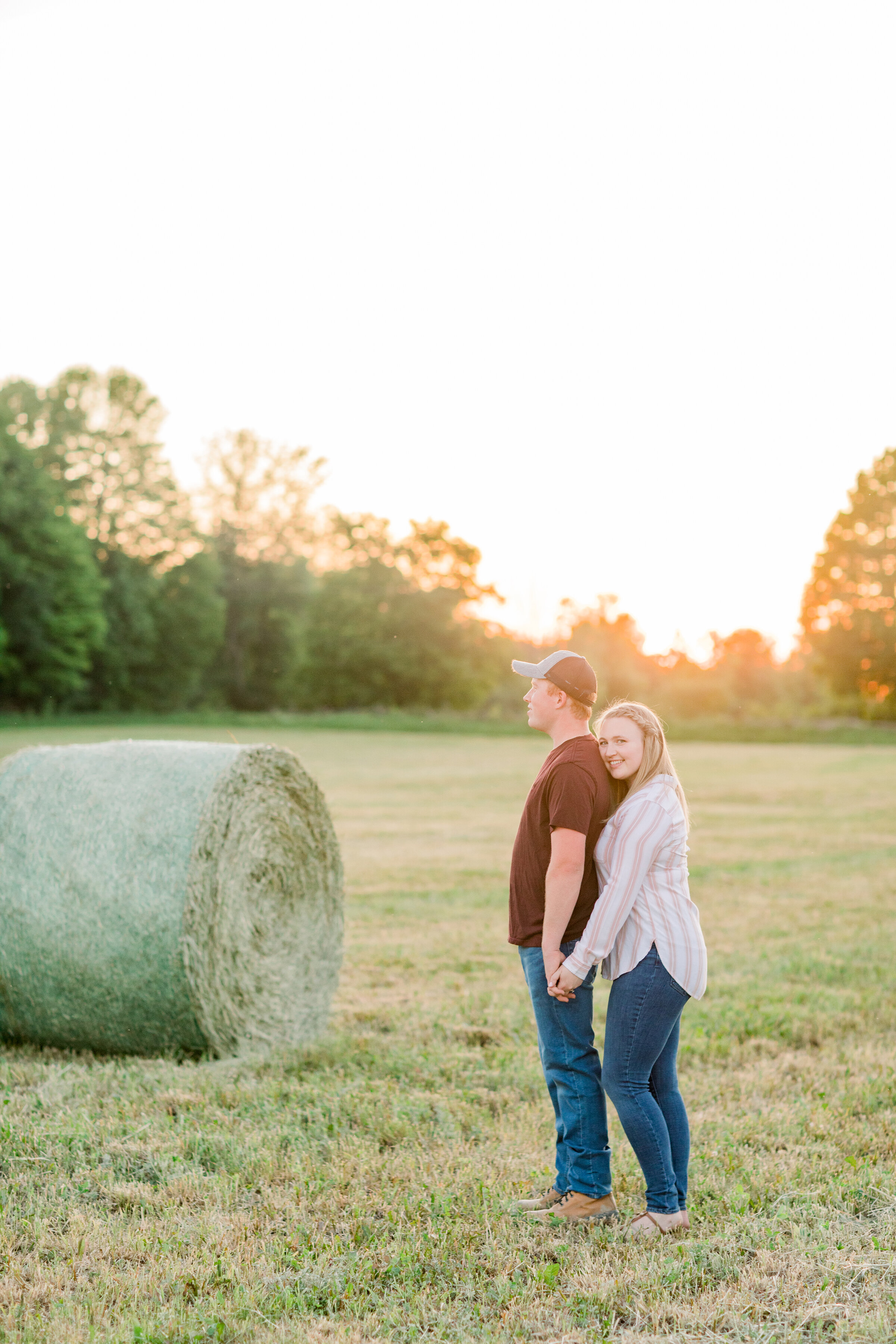  A couple stand together by a hay bail in a beautiful country farm themed engagement session by Chelsea Mason Photography. Engagement session goals couple goals couple pose inspiration glowing trees outdoor engagement session casual engagement attire
