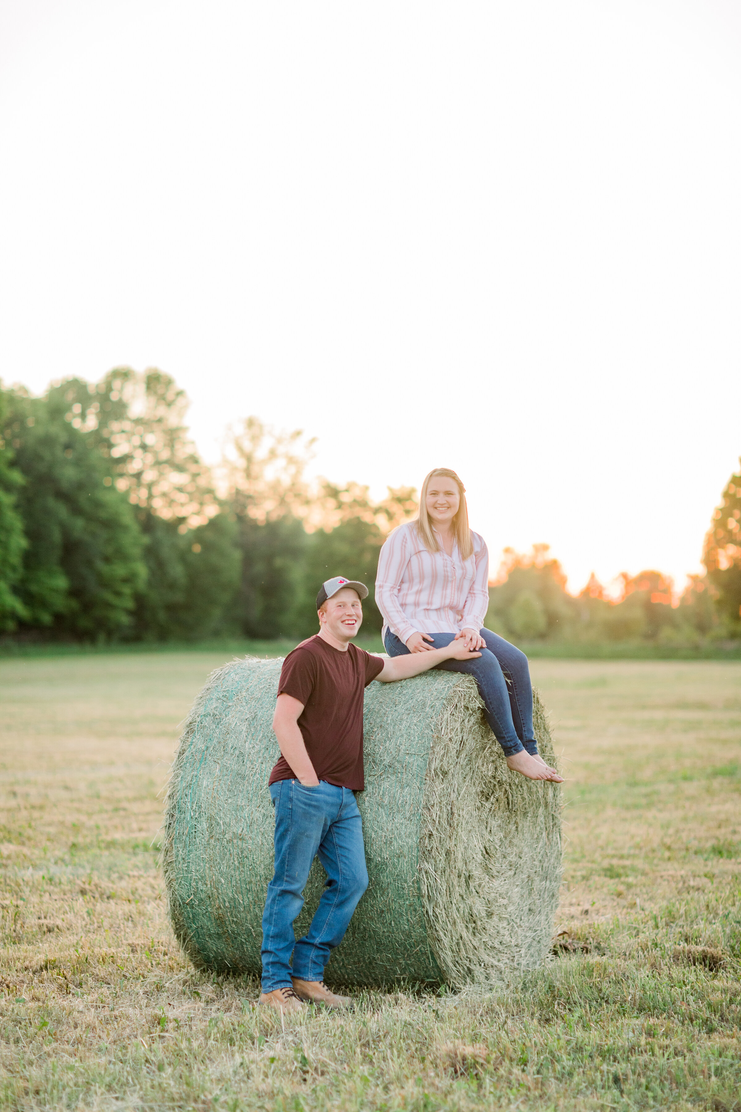  A  women sits on a hay bale while her husband stands beside her in a unique farm styled engaged session by Chelsea Mason Photography. Farm styled engagement session inspiration ideas and goals couple pose inspiration country life country styled outf